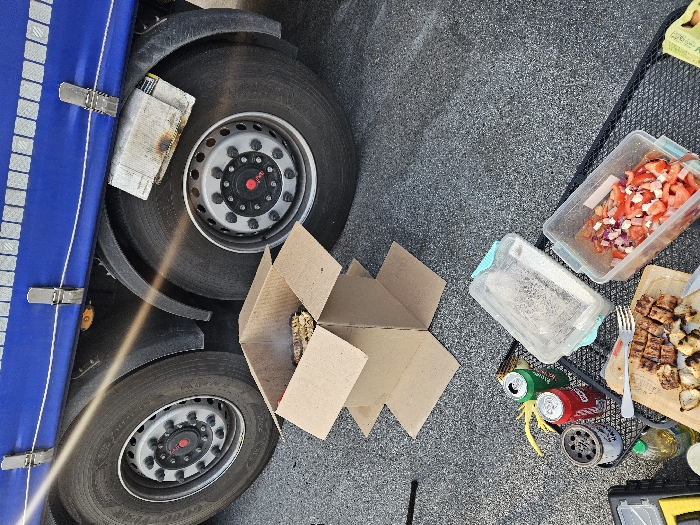 The image depicts an outdoor cooking and dining scene on dark grey asphalt pavement in Ventimiglia, Italy. The upper portion of the frame features the rear section of a commercial truck, characterized by a blue tarpaulin-covered trailer side secured by silver buckles, and two large black tires with silver hubcaps and red central caps. A black mudguard is positioned above the right tire, and a light-colored, folded fabric object secured above it displays burn marks on its lower edge. A bright sun flare diagonally crosses the upper left quadrant.
Centrally located is a makeshift grill fashioned from a brown cardboard box, containing visible glowing embers or charcoal and emitting smoke. Flat cardboard pieces form a base beneath this structure. The lower half of the image displays various items arranged on a black wire rack or directly on the ground. These include a wooden cutting board holding grilled, cubed meat (appearing to be chicken or pork) and some grilled onions, with a silver fork resting on it. Adjacent is a clear rectangular plastic container filled with chopped red tomatoes and white onion. Another larger, clear plastic container with a light blue lid is also visible. Two open aluminum beverage cans, one red and one green, are present. A small bottle of olive oil, a dark cylindrical object (possibly an ashtray or small burner), and a yellow tool are also discernible. A portion of a black object is visible on the far left. No individuals are present in the scene.