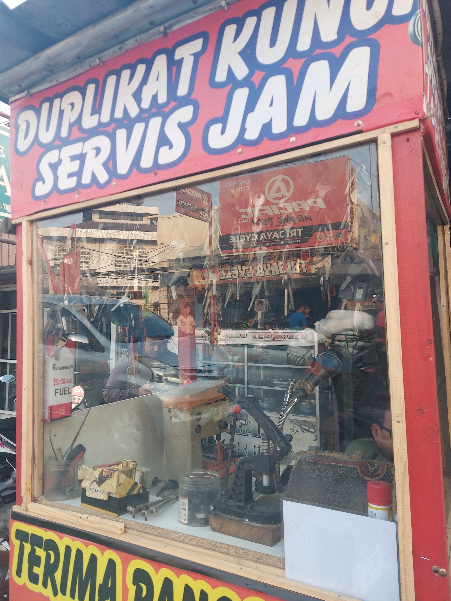 The image shows a storefront for a key duplication and watch repair service located in Karawang, Indonesia. The storefront features a red and blue sign with white lettering that reads "DUPLIKAT KUNCI" (Key Duplication) and "SERVIS JAM" (Watch Service). The front of the shop has a glass display window where various tools and equipment for key duplication and watch repair are visible, including a key cutting machine, pliers, and small containers of parts. Behind the glass, two individuals are present; one appears to be a customer looking at something on a table, and another person, presumably a worker, is behind a counter. In the background, reflections in the glass show a street scene with buildings and other shops. The overall impression is that of a small, local business operating in a busy urban environment.