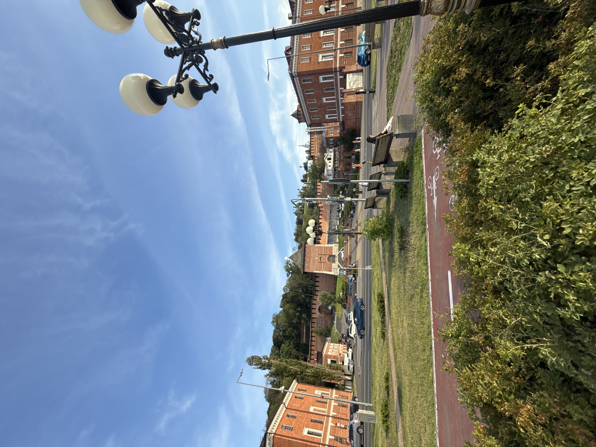 The image shows a bright, sunny day in Nizhny Novgorod, Russia. The scene is a panoramic view of a street with a red bike lane and a sidewalk. There are several brick buildings lining the street, some with multiple stories and visible windows. Green trees and bushes are abundant, adding to the natural scenery. A classic street lamp with multiple white globes stands prominently in the foreground, its ornate black metalwork detailed against the blue sky.
On the sidewalk, a person is relaxing on a wooden bench, looking away from the camera. There are other benches visible as well. A few cars are parked along the street and some are in motion, indicating moderate traffic. In the background, a large, ancient-looking brick fortress or castle structure is visible, perched on a hill surrounded by greenery. White domes of churches can be seen peeking out above the trees and fortress walls, suggesting a historic and culturally rich location.
The sky is a vibrant blue with thin, wispy clouds, indicating good weather. The lighting suggests it is daytime, likely late morning or afternoon, given the angle of the shadows and the brightness of the scene. The overall mood is peaceful and serene, with a hint of urban activity blended with historical architecture and natural beauty. No visible text is present in the image.
