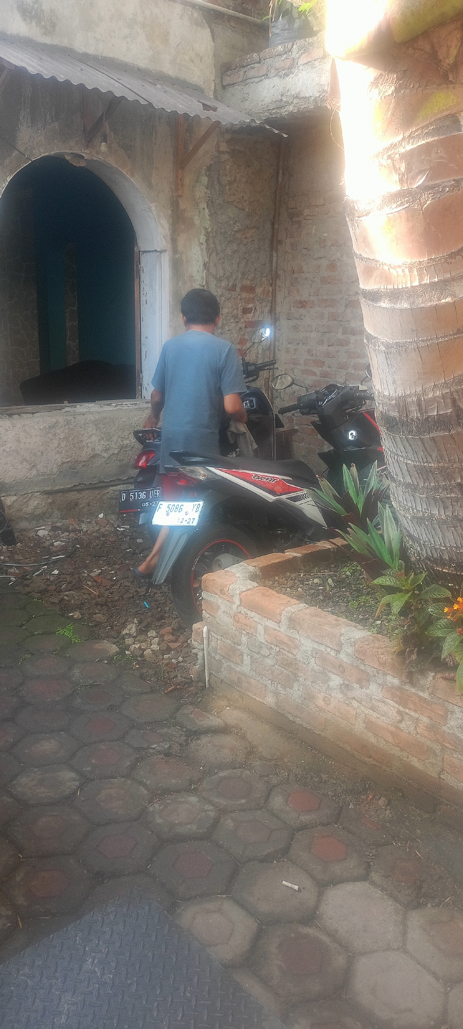 An unidentified male wearing a blue t-shirt and dark pants is standing next to a black motorcycle with red and white accents. The motorcycle has two license plates visible: one reads "D 5136 UER" and the other, "F 5086 YB" with "12-27" below it. The man appears to be holding a cloth in his right hand. The scene is outdoors, with a rough brick wall and a large tree trunk to the right. In the background, there is a building with an arched entrance covered by a corrugated metal awning. The ground is paved with hexagonal interlocking concrete blocks. The overall environment suggests a residential area in a city like Mekar Rahayu, Indonesia, with a slightly neglected or rustic aesthetic.