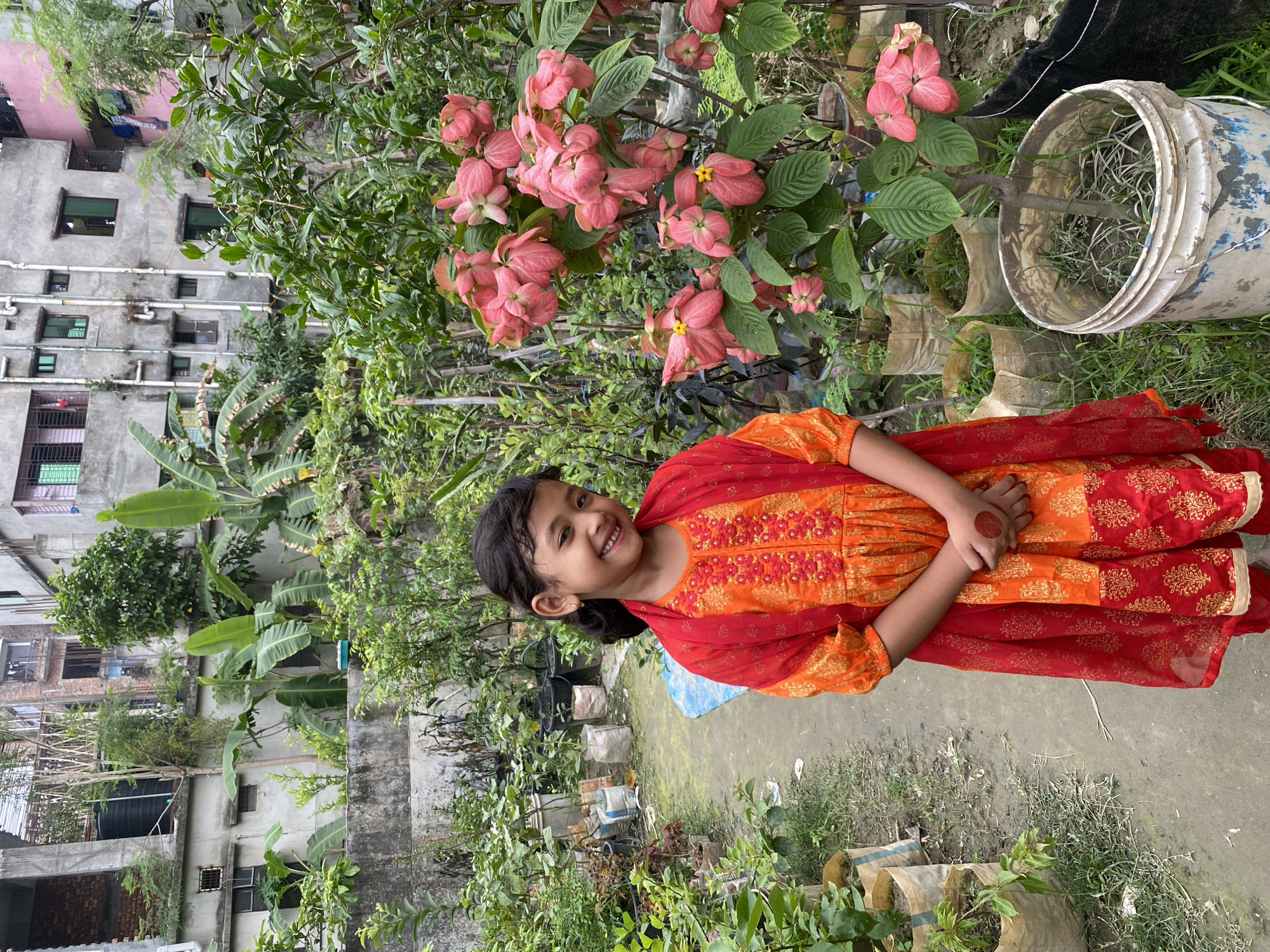 A young girl stands smiling in a lush garden area, likely a rooftop garden, with pink flowers behind her. She is wearing an orange and red dress. The background includes buildings and other greenery. The location is inferred to be Dhaka, Bangladesh, based on the architecture and vegetation. There are various potted plants and a white bucket visible in the garden. The path is adjacent to the girl, which appears to be concrete or pavement. The girl has a bindi on her forehead, suggesting cultural significance.