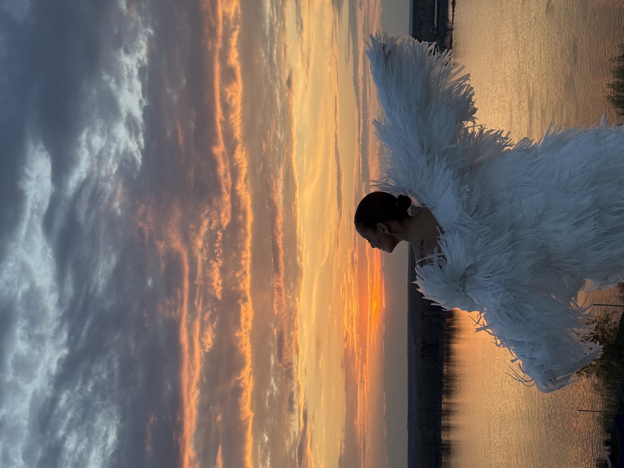 A rotated outdoor long shot shows a woman in a white feathery dress facing left, with a sunset and a body of water in the background. She is silhouetted against the light. The water reflects the golden hues of the sunset. The location appears to be near a body of water in or near the city of Ольховка, Russia, based on the presence of a sunset over water, common in areas with lakes or rivers. The woman seems to be posing, creating a serene yet striking image against the natural backdrop.