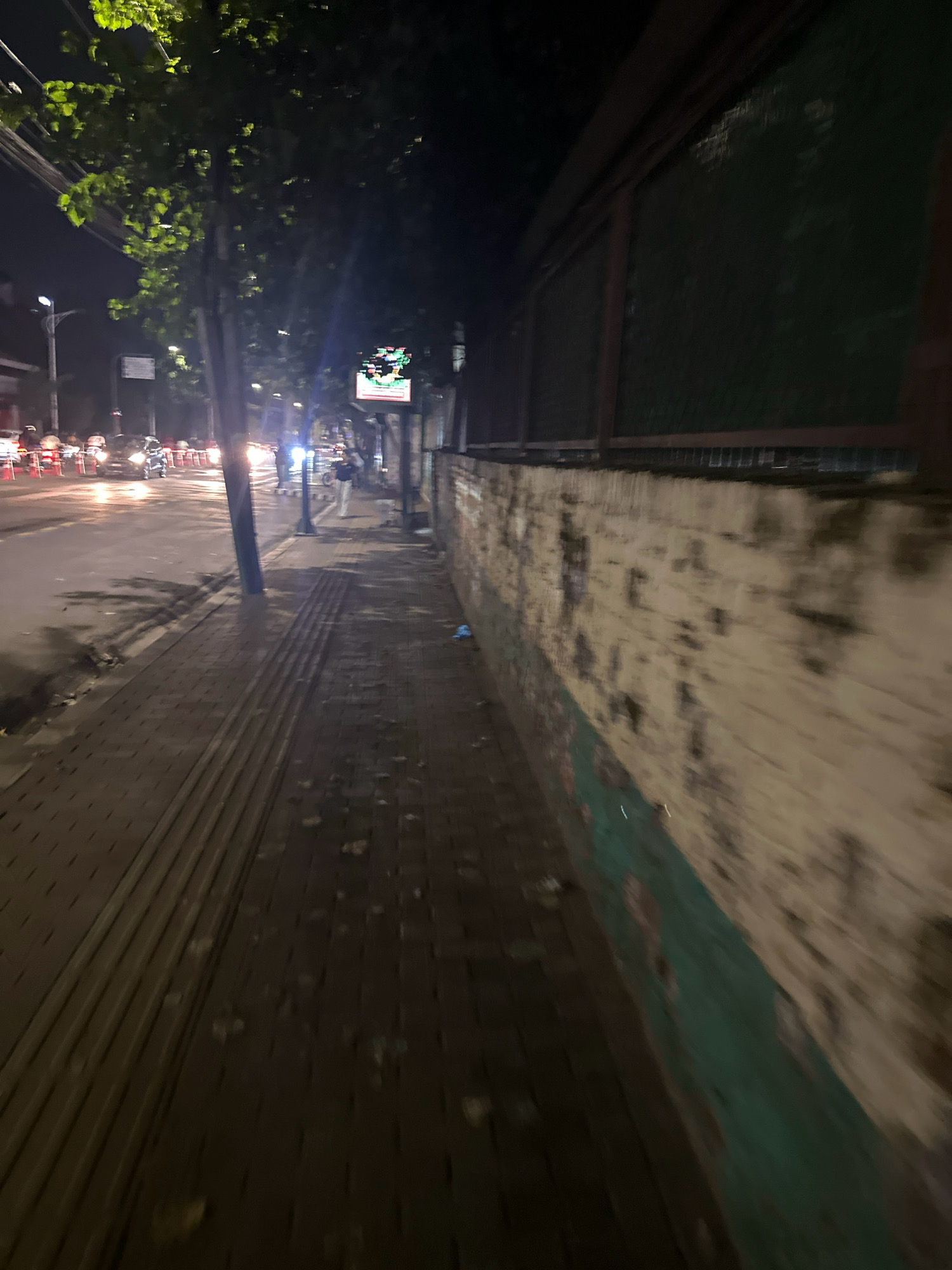 This image depicts a nighttime scene on a sidewalk in Kathmandu Metropolitan City, Nepal. The sidewalk is paved with dark bricks and features tactile paving for the visually impaired. A street lamp illuminates the scene, casting shadows. The street is wet, suggesting recent rain, and traffic lights and the headlights of vehicles are visible in the background. A tree with green leaves grows on the left side of the image, and a brick wall, partially covered in paint, runs along the right. A person in light-colored clothing is walking on the sidewalk. The overall atmosphere is urban and functional, with elements of typical street infrastructure.