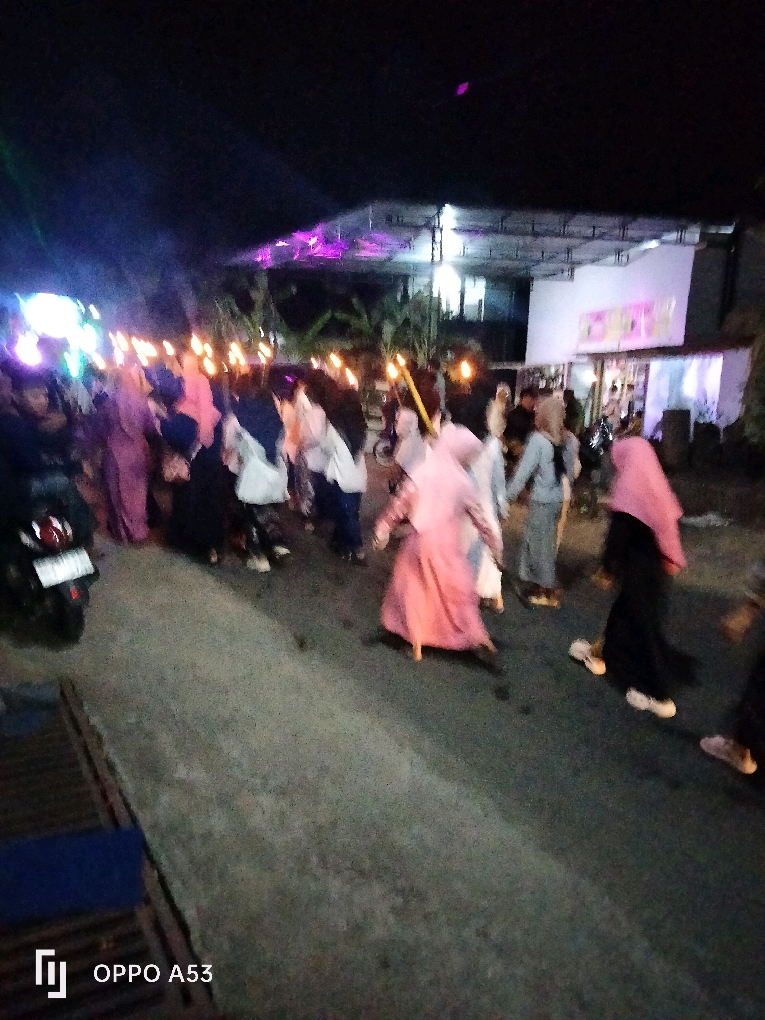 In Bendorejo, Indonesia, a nighttime street scene unfolds with a group of people holding torches, illuminated by their flames and artificial lights. The crowd, mostly women wearing hijabs and long dresses in various colors like pink, purple, black, and gray, are walking on a paved road. Some individuals are walking together, while others are more spread out. A motorcycle is parked on the left side of the frame, its license plate partially visible. In the background, a building with a white facade is lit with purple and white lights, suggesting a festive or ceremonial event. The overall atmosphere is dimly lit, with the torches providing a warm glow against the dark night sky.
