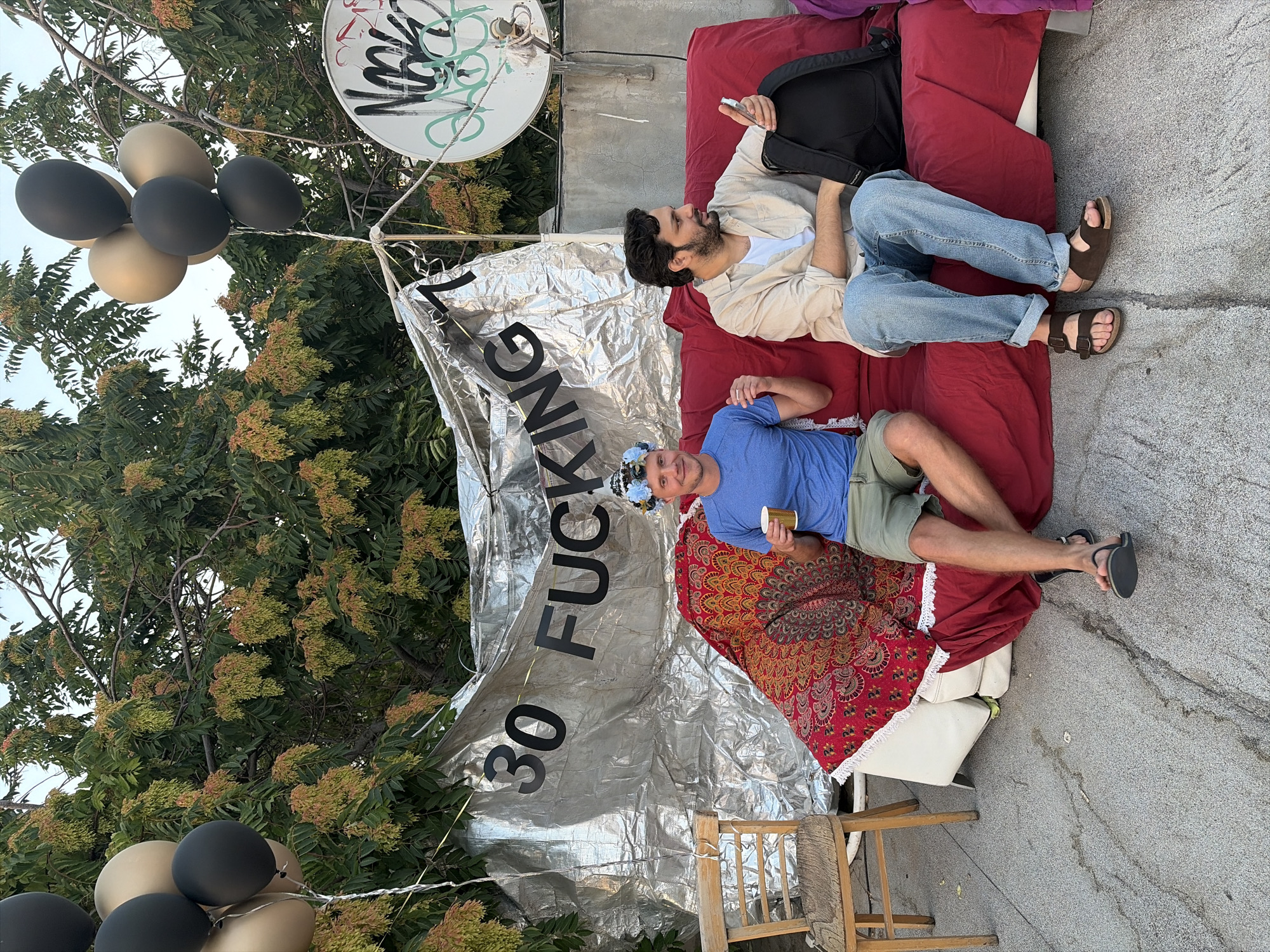 Two men are seated on a red couch in an outdoor urban setting. Behind them, a large silver banner with black lettering reads "30 FUCKING 7". Balloons are suspended from wires above the banner. The man on the left wears a blue t-shirt, green shorts, and a flower crown, and holds a cup. The man on the right wears a beige shirt, white t-shirt, blue jeans, and sandals, and is holding a smartphone. He has a backpack next to him. A wooden chair is to the left of the couch. In the background, there are trees, a satellite dish with graffiti, and a concrete wall. The ground is paved with grey gravel. This scene is located in Tel-Aviv, Israel.