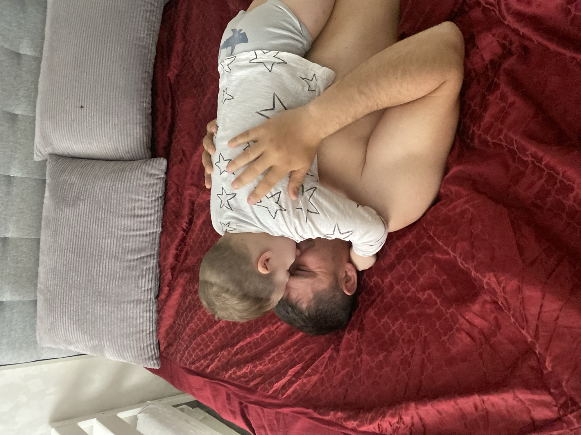 A top-down shot depicts a shirtless adult lying on a bed covered in a deep red blanket with a subtle repeating pattern. A young child, wearing a white shirt with black stars and light-colored shorts, is lying on top of the adult, face-to-face. The child's hand is visible on the adult's side. Two rectangular gray pillows are positioned to the left side of the bed. The location cannot be determined.