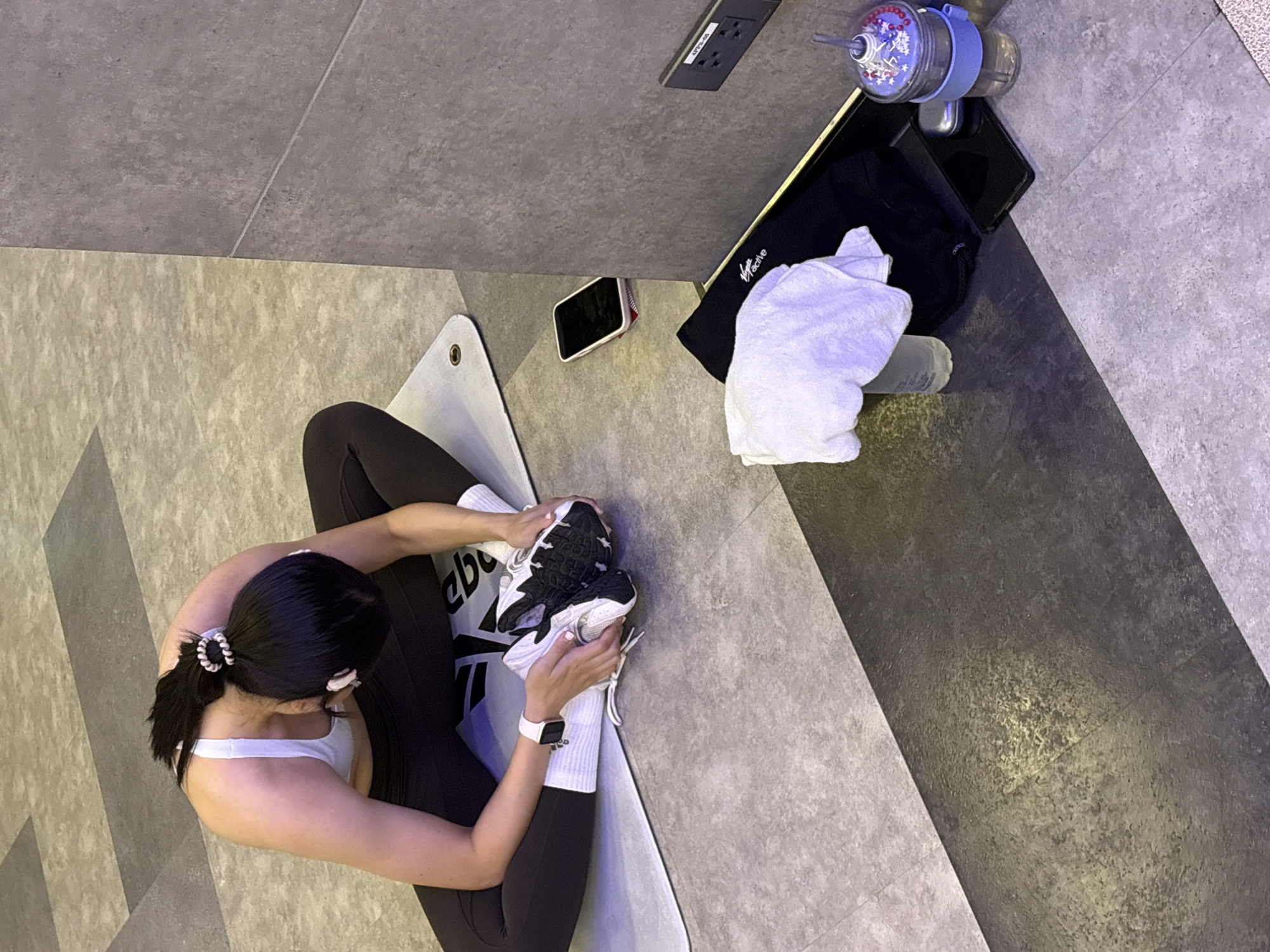 A woman is pictured from a top-down perspective, engaging in a yoga pose on a mat in what appears to be an indoor gym. She has dark hair pulled back with scrunchies and is wearing a white sports bra and black leggings. Her right foot is flexed and held by her hands as she stretches her hamstring. She has a smartwatch on her left wrist.
Next to the yoga mat, on a textured grey surface, are several objects. These include a black gym bag with white lettering that appears to say "Reebok", a white towel, a dark-colored smartphone, and a blue and clear water bottle with a decorative lid. An electrical outlet is visible on the wall behind these items.
The floor is tiled in shades of grey, with darker grey rectangular sections interspersed with lighter grey. The lighting suggests an indoor setting, possibly during the day. The woman's posture and focus indicate a moment of quiet concentration during her exercise routine. The overall scene conveys a sense of personal wellness and a typical gym environment.