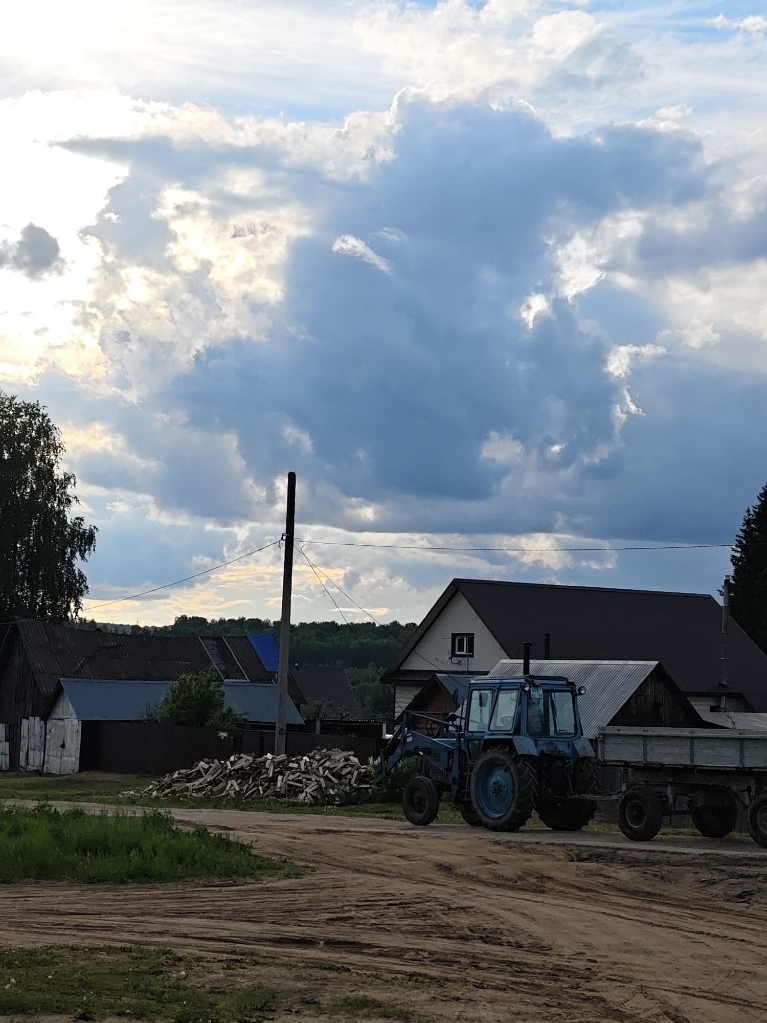 The image depicts a rural scene in Уральское, Russia. A blue tractor with an attached trailer is positioned on a dirt road in the foreground. Behind the tractor, there's a pile of chopped wood and a collection of various houses and structures. Overhead utility lines stretch across the sky. Above these features, the sky is filled with a mix of light and dark clouds. A few trees frame the scene on either side.