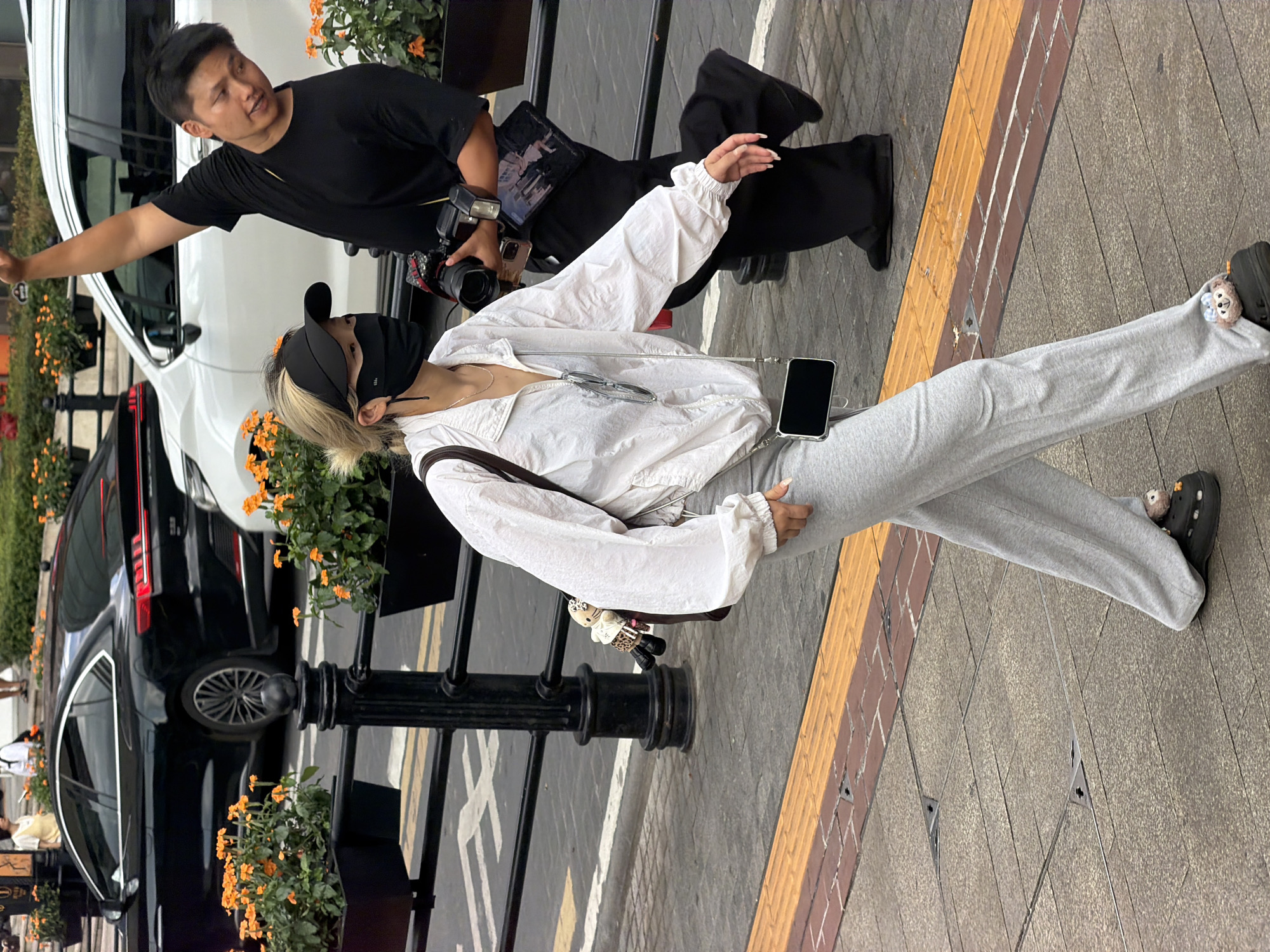 In Yuzhong District, China, a woman walks on a sidewalk with a black face mask, a black visor, and a white zip-up jacket, carrying a bag with a stuffed animal charm. She wears grey sweatpants and black Crocs with charms. Behind her, a man in a black t-shirt holds a camera and a phone. They are near parked cars and planters with orange flowers, with a black metal fence in the background. The sidewalk has textured tiles and a yellow tactile warning strip.