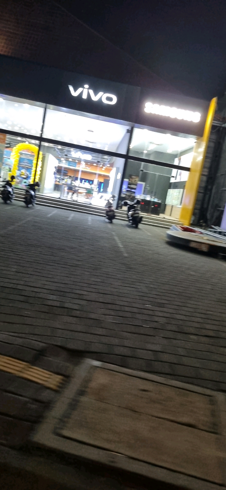 The image shows the exterior of a Vivo and Samsung store at night in Denpasar, Indonesia. The store has large glass windows and is brightly lit from within. The Vivo logo is prominently displayed on the facade. Outside the store, there are several motorcycles parked on a wooden boardwalk. The ground is paved with dark wooden planks. In the background, a large yellow vertical structure is visible, possibly a part of the building's architecture or a sign. The scene is partially obscured by motion blur, suggesting the photo was taken while in motion.
