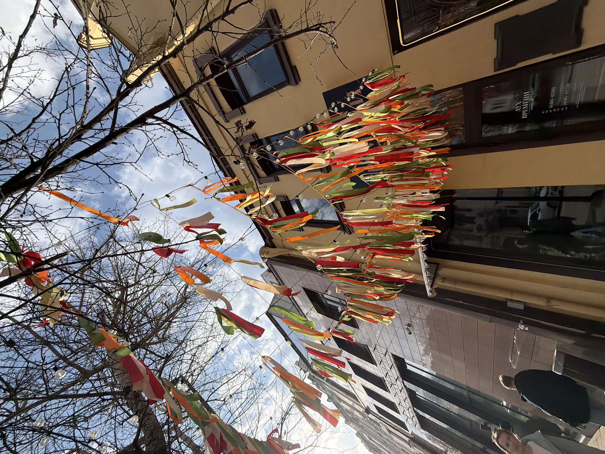 The image, taken in Krasnoyarsk, Russia, shows a low-angle view of a city street. Overhead, colorful ribbons are strung between buildings and trees, creating a festive canopy. The ribbons are primarily red, orange, green, and white.
A bare tree branches into the lower left of the frame, its silhouette contrasting against a cloudy, blue sky. The surrounding buildings are multi-storied with varied architectural styles, including modern glass structures and traditional facades.
In the lower right corner, two individuals are partially visible. One is wearing a black coat and appears to be walking away from the camera. The other is cut off by the frame but seems to be looking up at the ribbons.
