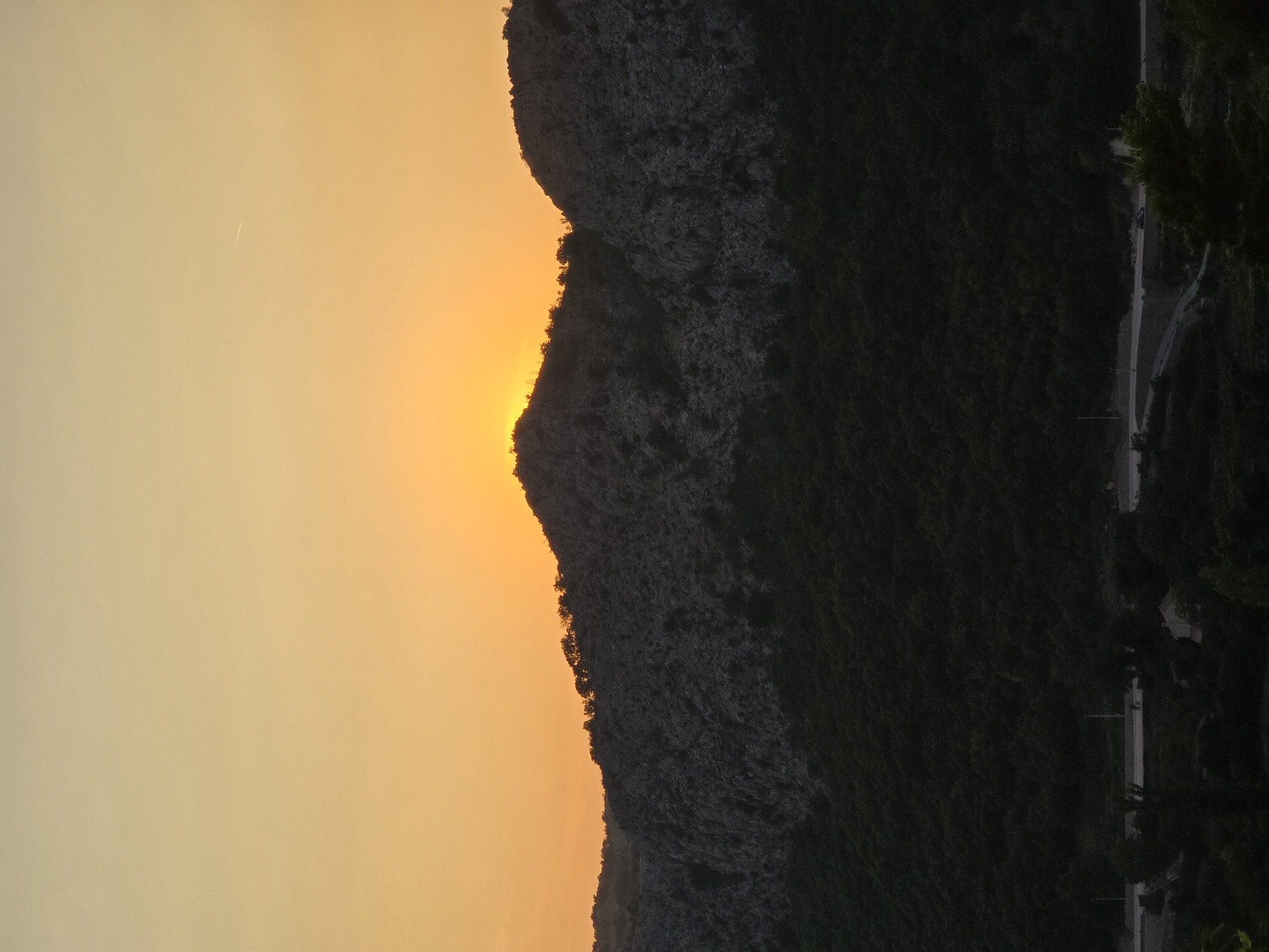 The image captures a sunset over a mountainous landscape near Gornja Vrbica, Montenegro. The sky is a warm gradient of orange and yellow, with the sun just peeking over the crest of a dark, rocky mountain. The mountain range is covered in dense green foliage in its lower half, transitioning to a rugged, grey, and stony texture higher up. The peaks are silhouetted against the bright sky, with a scattering of trees along the ridge line. Below the mountains, a dark, forested area is visible, obscuring the ground level. A road can be faintly seen winding through the lower part of the image, with a small building and some trees nearby. The overall scene is one of natural beauty and tranquility, characteristic of a rural or mountainous setting during twilight.