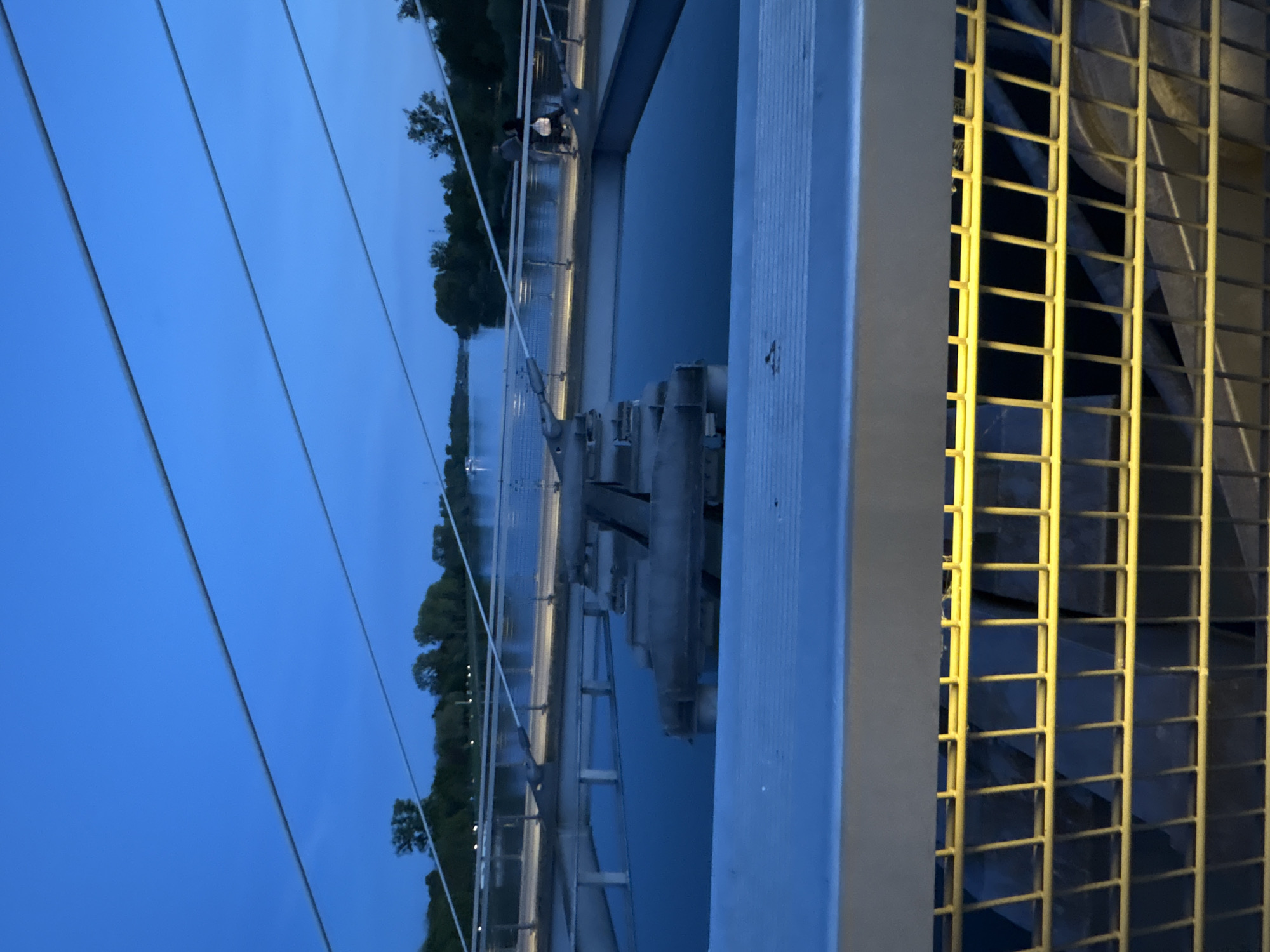 This image captures a tranquil twilight scene of a bridge spanning a wide body of water, likely a river or canal in Strasbourg, France. The sky is a deep, clear blue, indicative of dusk or early night, with dark silhouettes of trees lining the distant banks.
In the foreground, the robust structure of a bridge is prominent, featuring light-colored railings and several taut cables stretching diagonally across the frame, suggesting a cable-stayed design. To the right, a distinctive bright yellow metal grid or grating is illuminated, possibly part of the bridge's infrastructure or an adjacent mechanical section.
A lone individual, dressed in dark clothing, is visible on the bridge structure in the mid-ground, appearing to walk or stand. Beyond this figure, a series of complex structures extend across the water, possibly forming part of a lock system or a dam, with various mechanical elements and platforms. Distant lights from the banks and potentially from the bridge itself punctuate the scene, casting subtle reflections on the calm water surface. The overall mood is serene and peaceful. No text is visibly present in the image.