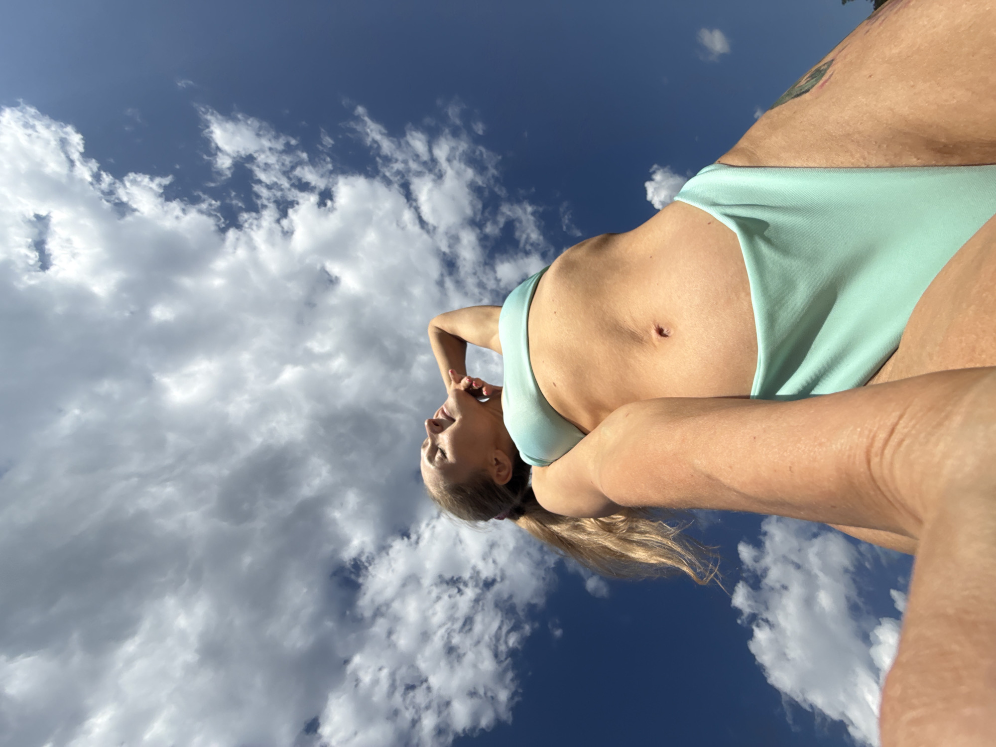 A woman in a light blue bikini is positioned against a background of blue sky and scattered white clouds. The camera angle is low, looking up at the woman. The location is not identifiable as Chaikovsky, Russia.