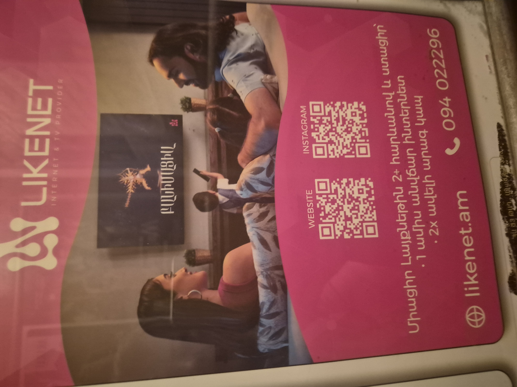 The image displays a pink promotional card for "LIKENET INTERNET & TV PROVIDER," an Armenian company. The card is angled and shown vertically, covering part of a larger background image.
On the card, the company's logo and name "LIKENET" are visible in white on the left. The right side features Armenian text detailing a promotional offer: "Join Likenet with 2+ neighbors and get: • 1 month free internet • 2x faster connection." Below the offer, the website "likenet.am" and a phone number "094 022296" are printed. Two QR codes are present, labeled "WEBSITE" and "INSTAGRAM."
The background image, partially obscured by the card, shows a family in what appears to be a cozy living room. A man with long dark hair and a beard, wearing a light shirt, is seated on a sofa, looking towards a child whose head is partially visible. A woman with dark hair, wearing a pink top, is lounging on an adjacent part of the sofa, looking upwards. The family appears relaxed and comfortable. A screen or picture frame on the wall behind them displays the Armenian word "ԲԱՆԻՄԱՑԻԱ" (Logic/Rationality). A small potted plant sits on a side table. The scene evokes a sense of domestic tranquility and family bonding, likely aiming to associate Likenet's services with comfort and home entertainment. The overall impression is one of a warm, inviting home environment. The use of Armenian language on the card and background element indicates its context within Armenia, such as Yerevan.
