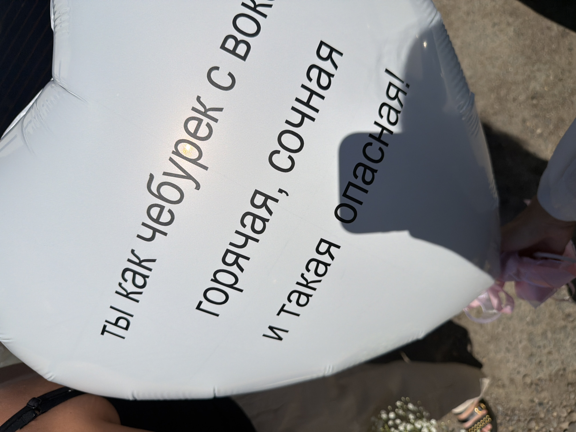 The image shows a white, heart-shaped balloon with black text in Russian. The text reads "Ты как чебурек с вокзала, горячая, сочная и такая опасная!" which translates to "You are like a cheburek from the train station, hot, juicy, and so dangerous!". A person is holding the balloon. Another person's hand holding a pink face mask is visible. A woman's shoulder with a black bra strap is on the bottom left. There are flowers and part of a sandal visible as well. The ground is concrete. Based on the Cyrillic text and the reference to "вокзала" (train station), and its proximity to Chaikovsky, Russia, a reasonable hypothesis is that the location is somewhere in or near Chaikovsky.