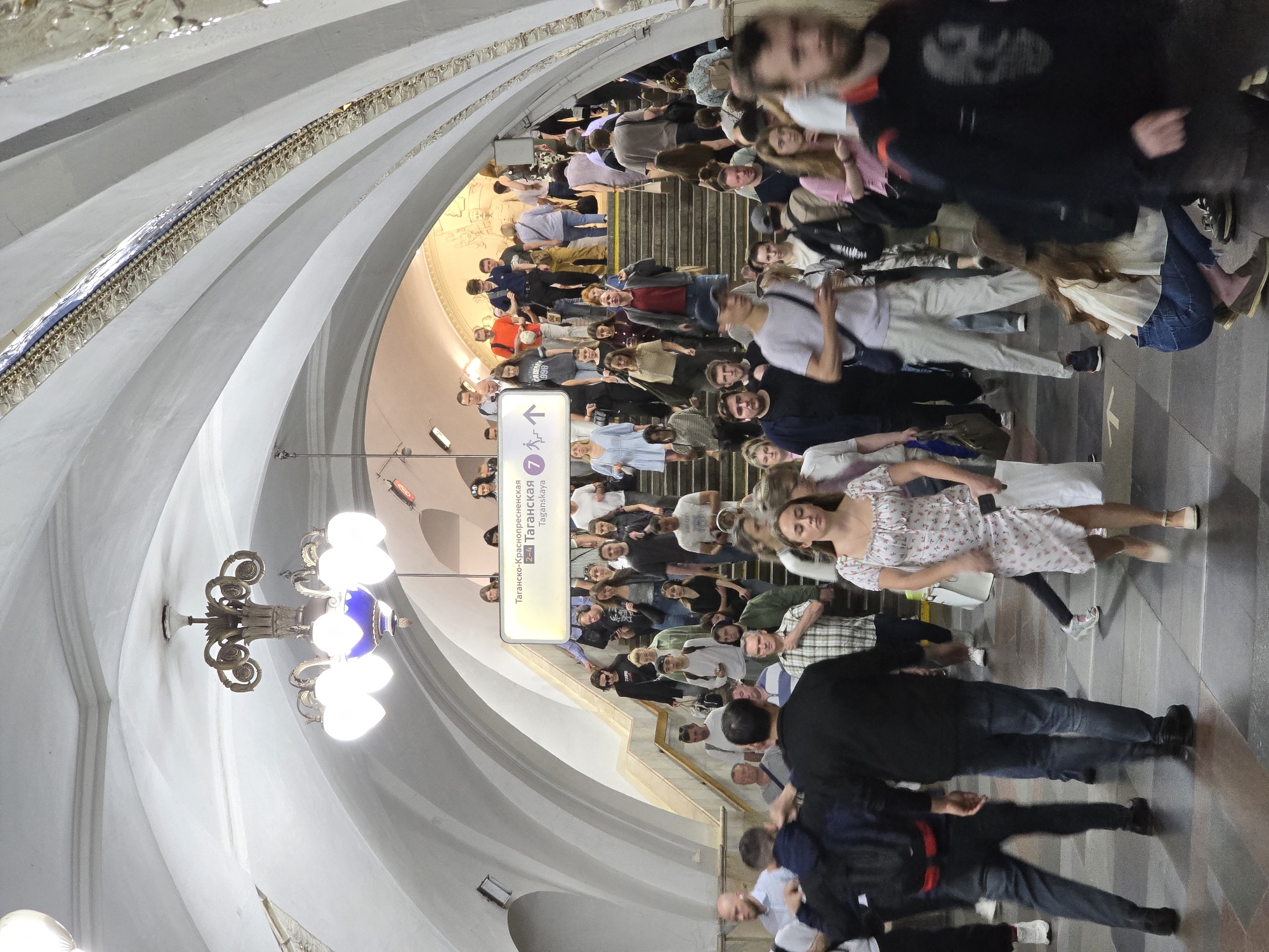 The image captures a scene inside what appears to be a subway station. A large number of people are ascending a set of stairs. Overhead, there is a decorative chandelier and a sign indicating "Taganskaya" in Cyrillic, likely pointing towards the Taganskaya station in Moscow, Russia. The architecture features arched ceilings with ornate detailing along the edges. The scene conveys a sense of crowdedness and movement within the station.