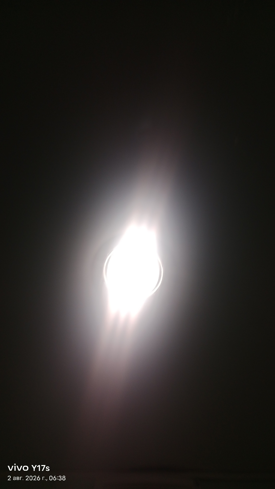 The image shows a bright, possibly incandescent, light source emitting light in a dark, possibly dusty or smoky, environment. The light source is centrally located and appears to be a bulb with a reflective or frosted outer shell. There are lens flares or light streaks emanating from the bulb, extending upwards and outwards. The background is dark and underexposed, with a gradient from dark grey to black. In the bottom left corner, text overlay indicates "vivo Y17s" and the date "2 авг. 2026 г., 06:38", suggesting the photo was taken with a Vivo Y17s phone on August 2, 2026, at 06:38 AM. The location is specified as Rudny, Kazakhstan.