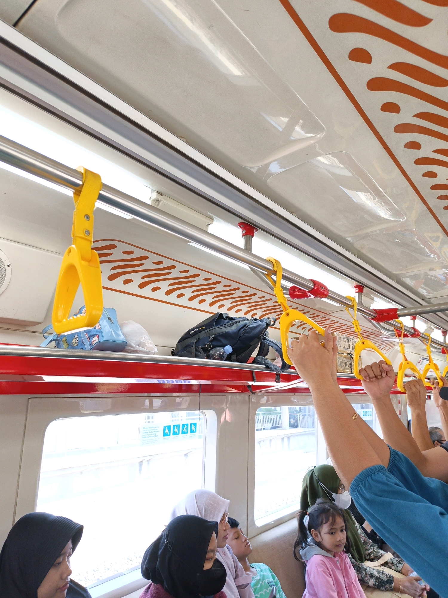 The image depicts the interior of a commuter train, likely in Delanggu, Indonesia, focusing on the passenger area. Several yellow grab handles are visible, attached to metal poles running along the ceiling, some with red accents. Bags, including a blue tote and a black backpack, are stowed on a shelf above the seats. Passengers are seated and standing, with some holding onto the grab handles. Two individuals, presumably passengers, are seen from the chest up with their arms raised, holding onto the yellow handles. Their attire suggests a modest style, with headscarves visible on several women. A young girl in a pink jacket is seated in the foreground, looking down. The train windows show a blurred outdoor scene, indicative of movement. The interior design includes a patterned orange graphic on the ceiling and red trim along the window frames. The lighting appears to be a mix of overhead interior lights and natural light coming through the windows.