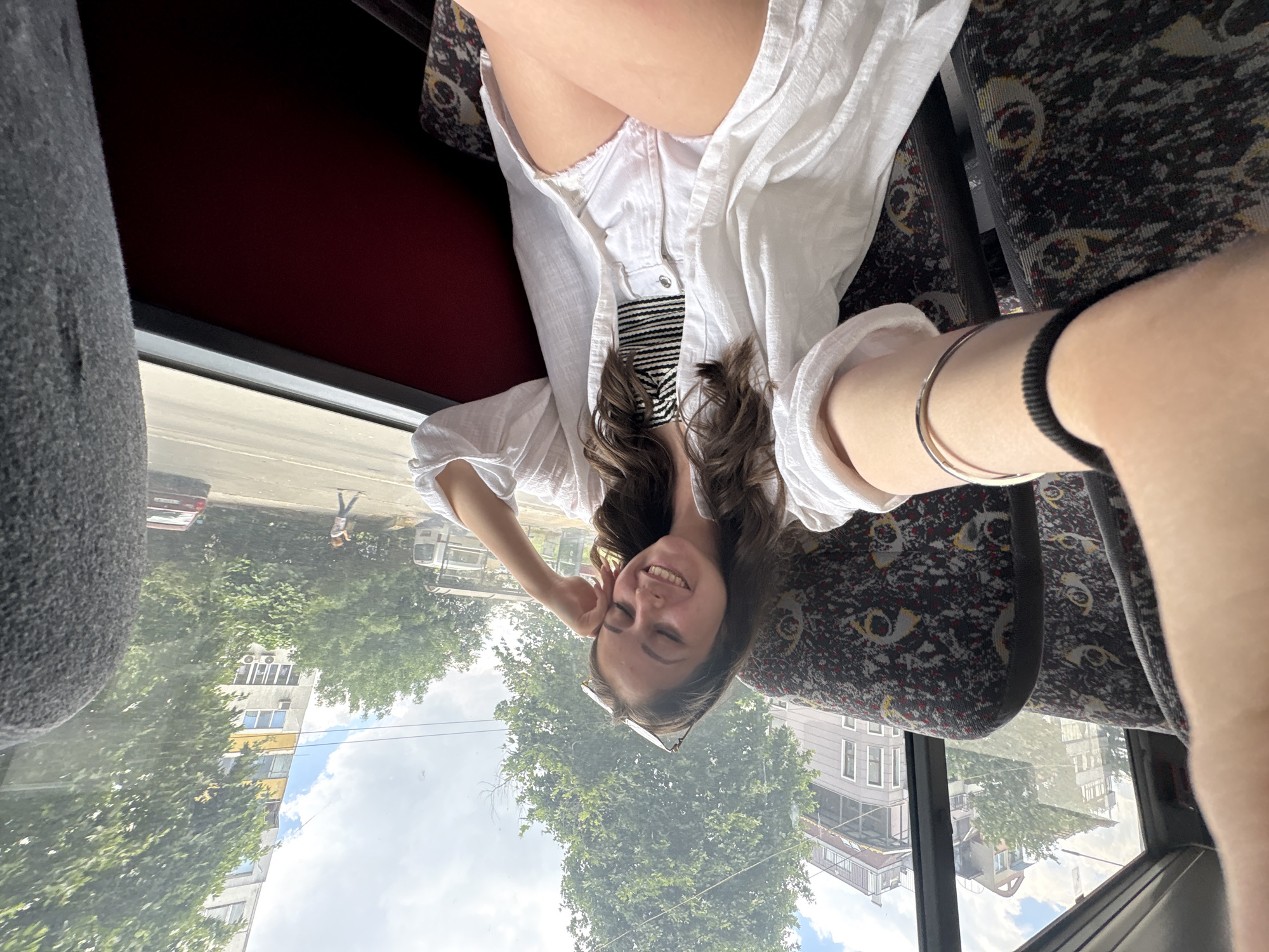 A young woman with long, dark, wavy hair, wearing a white open-necked shirt, a black and white striped top, and white denim shorts, smiles with her eyes closed while sitting on a bus in Burgas, Bulgaria. She has sunglasses perched on her head and her left arm rests on her cheek. The bus seats are upholstered in a patterned fabric. Through the window, apartment buildings and trees are visible, suggesting an urban environment. The weather appears to be partly cloudy.