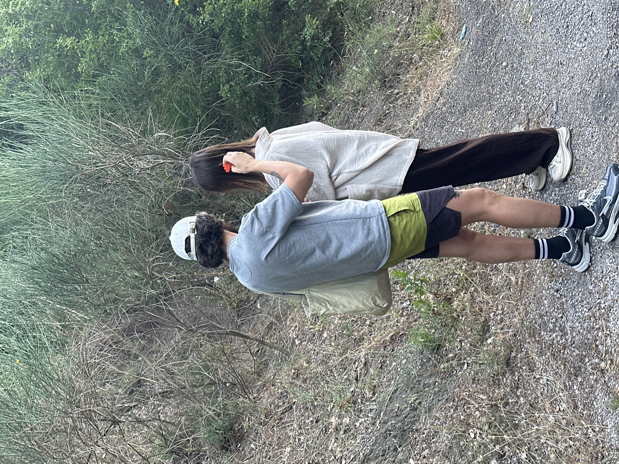 The image shows two individuals, one wearing a grey t-shirt and lime green shorts, and the other wearing a cream-colored oversized shirt and dark brown pants. The person in the t-shirt has a canvas tote bag slung over their shoulder and is wearing a white baseball cap. The person in the cream shirt has a red object, possibly a flower, being placed behind their ear by the other individual. They are standing on a gravel path next to a grassy, overgrown embankment. The environment appears to be a natural, rural setting, likely in Čučuci, Montenegro, characterized by dry vegetation and dense green foliage in the background. The lighting suggests it is daytime.