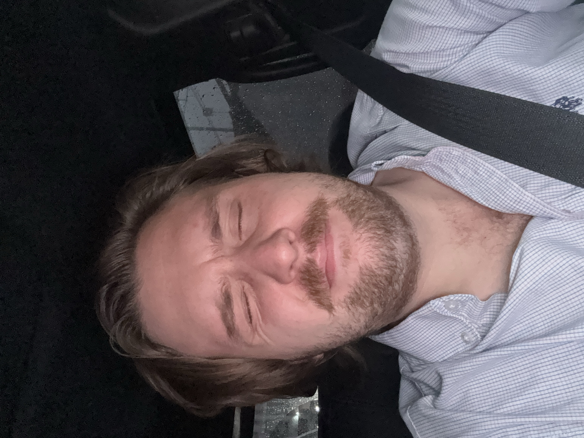 A man is in a car, wearing a light blue striped shirt and a seatbelt. His eyes are closed. The car appears to be in Mexico City, Mexico, as there is evidence of raindrops in the window.