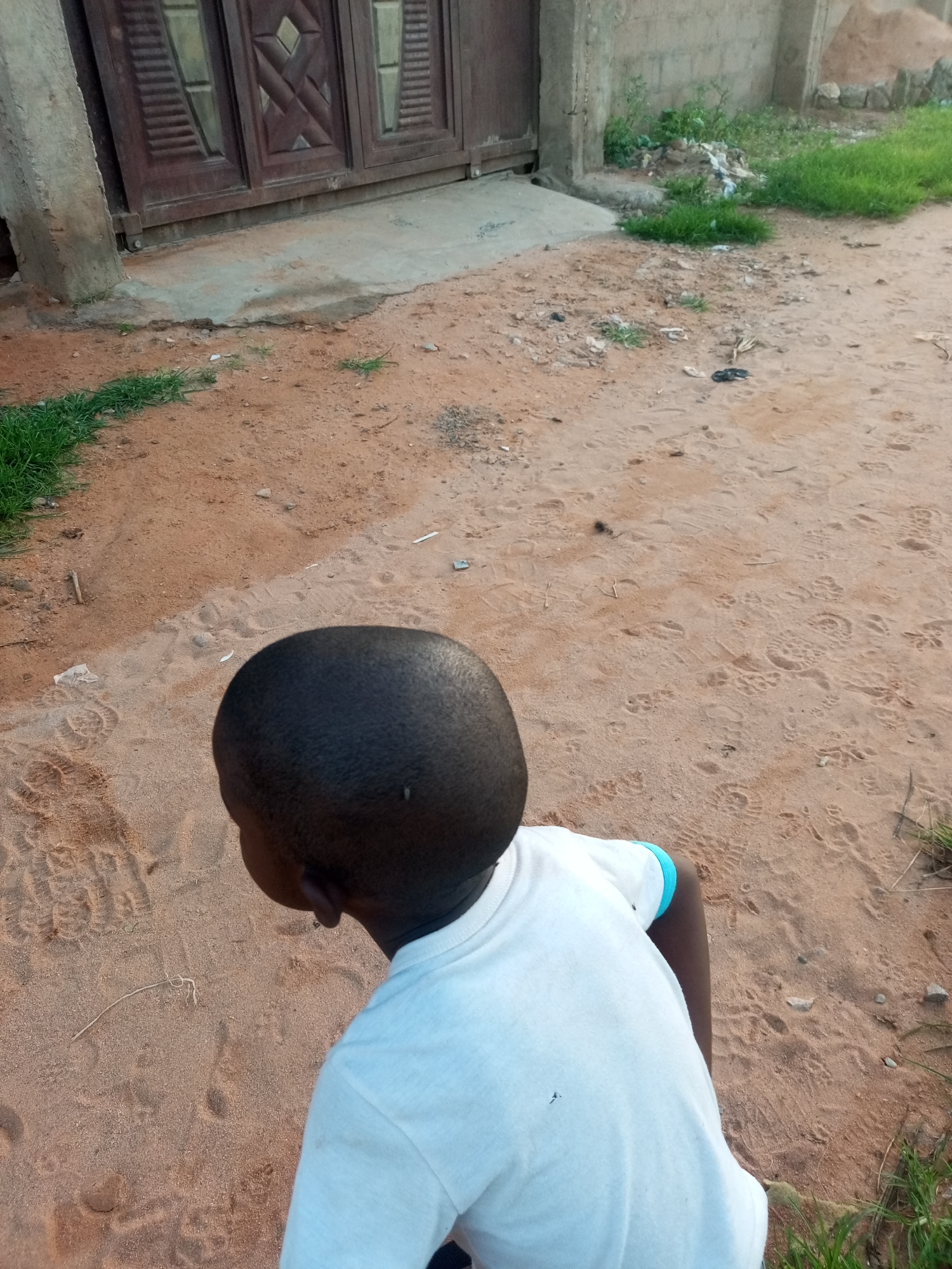 An overhead shot shows a young boy from the shoulders up, facing away from the camera. He has dark skin and a close-cropped haircut. He is wearing a white t-shirt with a blue band around the left sleeve. The boy is standing in sandy soil that has many footprints in it. To the left of the boy is a concrete pillar and a large, dark brown metal gate with geometric patterns. Behind the gate is a building. To the right of the boy, the sand gives way to patches of green grass and some scattered debris. The scene appears to be outdoors in a dusty area, possibly a residential street or yard.