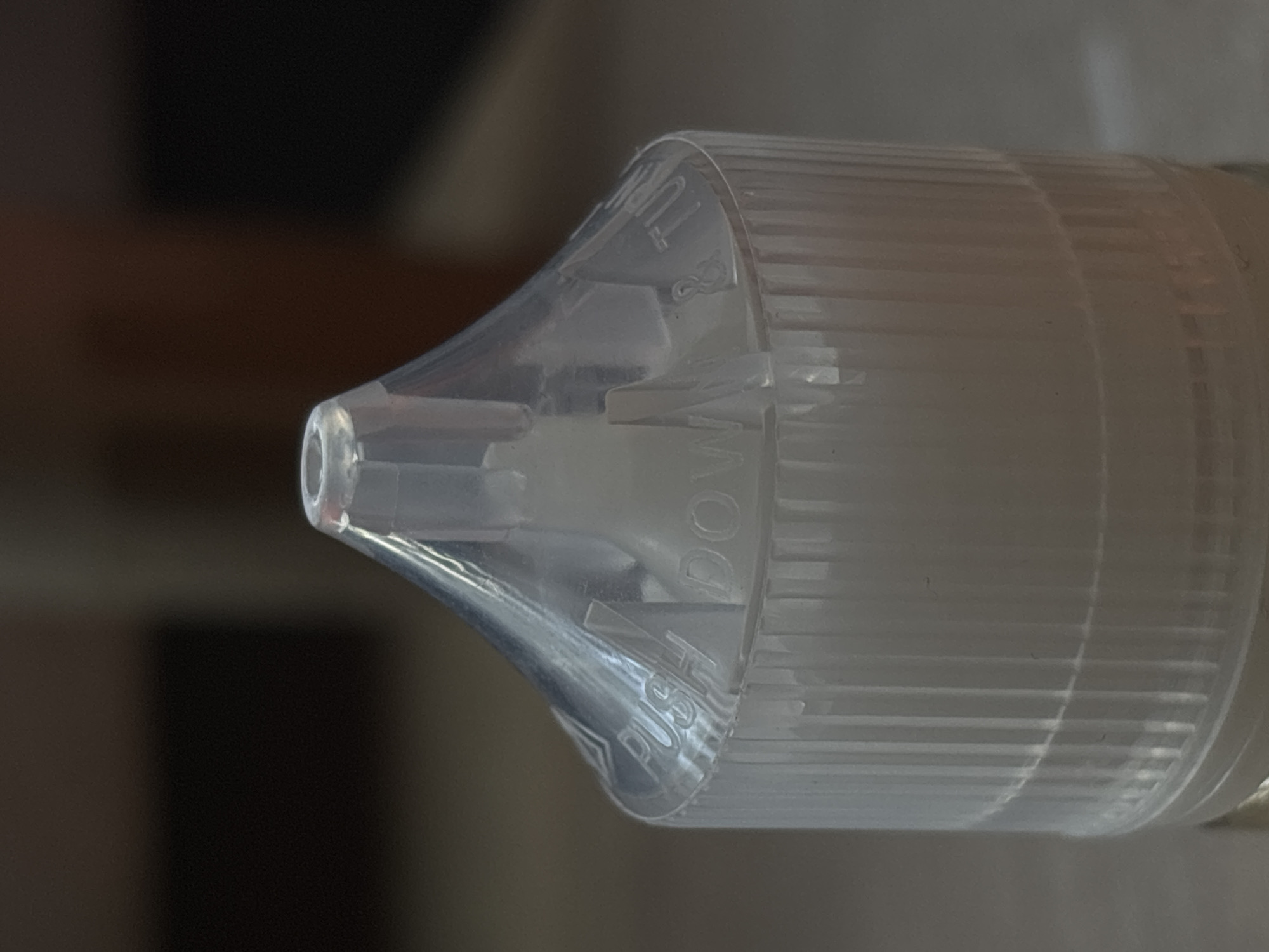 The image shows a close-up of the cap of a clear plastic bottle, likely for e-liquid or similar product. The cap has a conical shape with ribbed sides and a nozzle at the top. Raised lettering on the cap reads "PUSH DOWN & TURN". The background is blurred and dark, suggesting an indoor setting. There are no people or specific landmarks visible that would indicate a location in Saint Petersburg, Russia. The lighting appears to be artificial and focused on the bottle cap.