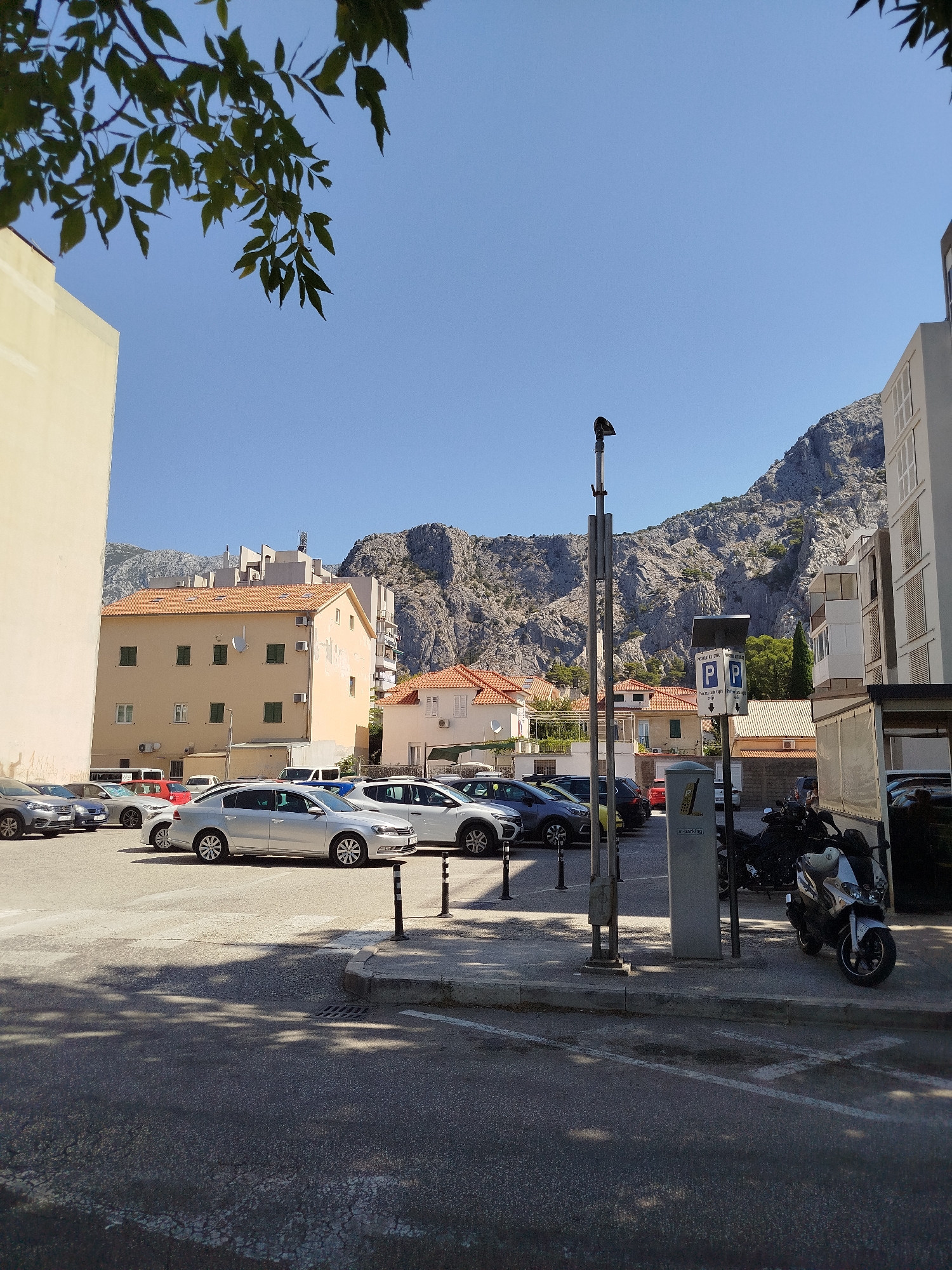 A street scene in Omiš, Croatia, features a parking lot filled with various cars and a motorcycle. The scene is dominated by a large, rugged mountain in the background under a clear blue sky. Several multi-story buildings with tiled roofs are visible in the middle ground. A modern, light-colored building with windows is on the right side of the image, and a large, light-colored building with a plain facade is on the left. A parking payment machine is situated next to a pole with a light fixture. Tree branches with green leaves hang from the top left corner of the image, casting shadows on the ground.