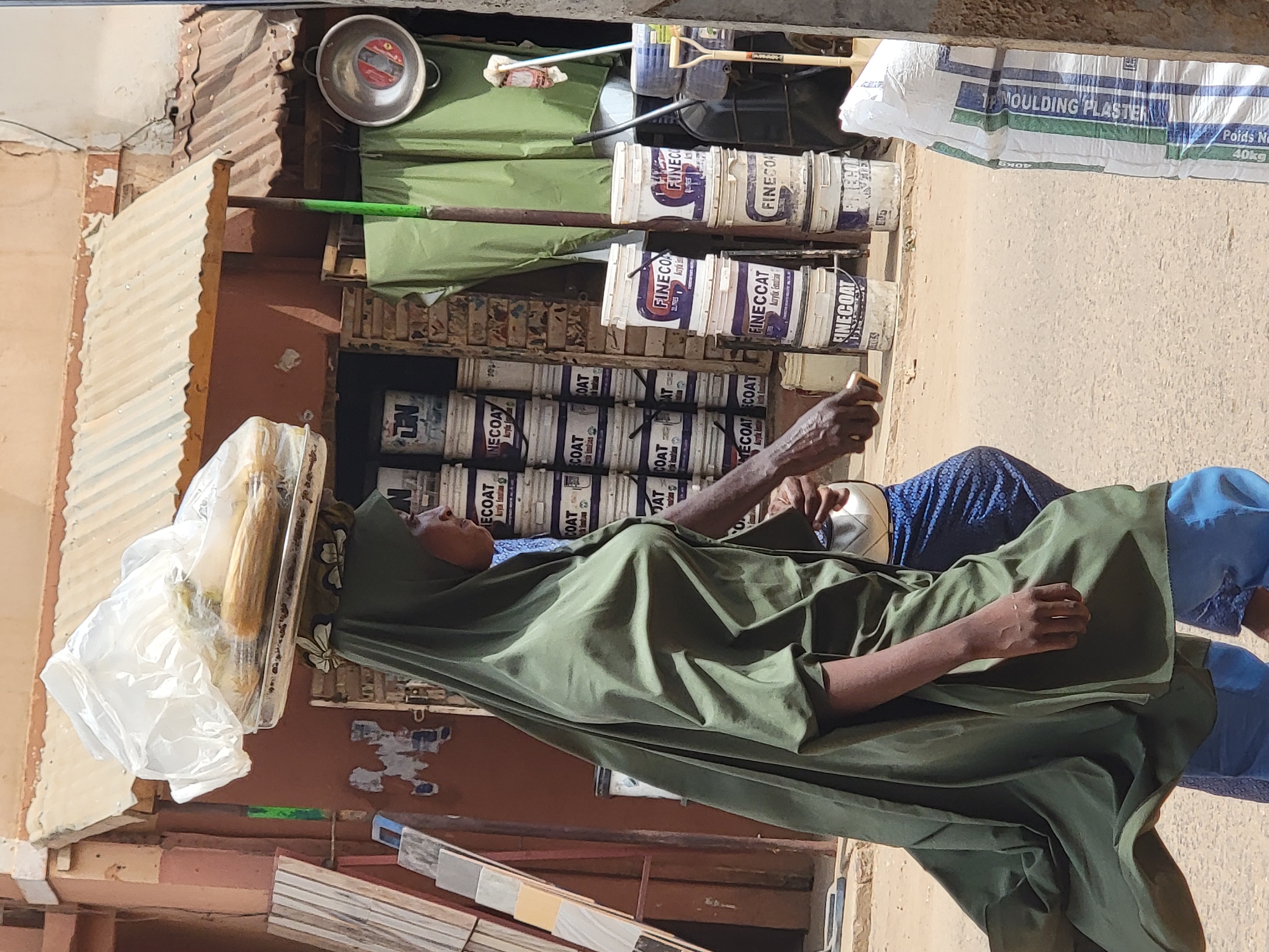 A woman in Maiburiji, Nigeria, is seen carrying goods on her head in front of a store. The woman is wearing a green head covering and robe. A clear plastic bag containing bread or other items is balanced on her head. The store behind her sells paint and other building materials, including buckets of "FINECOAT" paint and a bag of "MOULDING PLASTER." A metal bowl is also visible on top of the store's shelving. The weather appears to be sunny and bright.