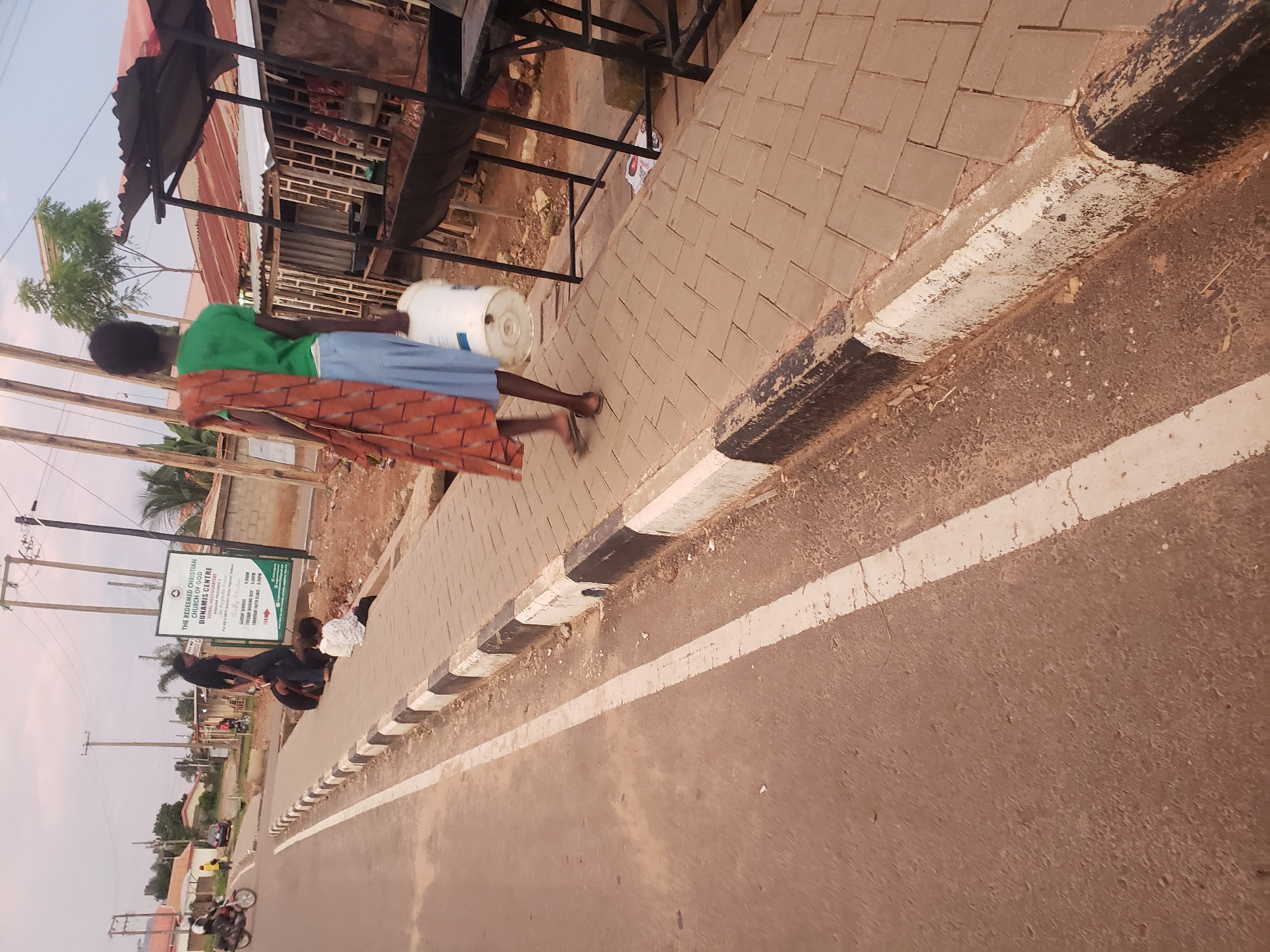 The image depicts a street scene in Narayi, Nigeria, with a focus on a pedestrian walkway adjacent to a road. In the foreground, a young person is walking along the paved sidewalk, carrying a white bucket and draped in a patterned cloth. The individual is wearing a green shirt and blue skirt. Two children are seated on the sidewalk near a roadside sign. The sign, in English, identifies "THE REDEEMED CHRISTIAN CHURCH OF GOD DUNAMIS CENTRE" and provides service times and contact information. A motorcycle is visible in the background on the road, and several structures, including market stalls with corrugated metal roofs, line the opposite side of the street. The environment appears to be a bustling urban area with signs of daily commerce and community life.