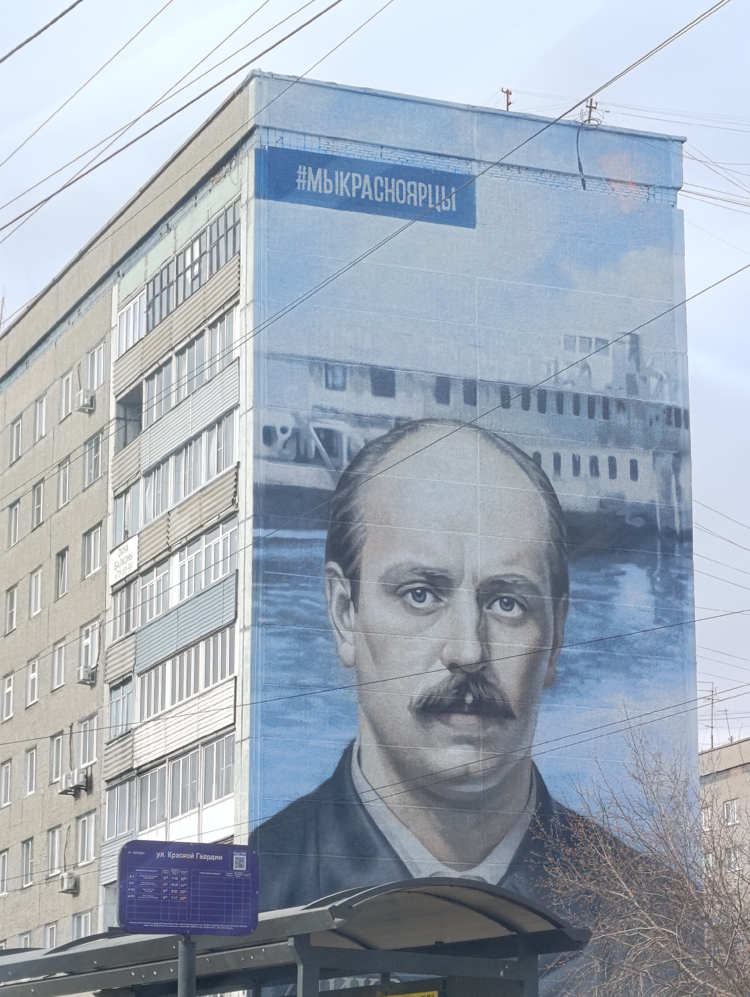 This image presents an urban street scene in Krasnoyarsk, Russia, featuring a multi-story residential building under a bright, overcast sky. The dominant element is a large mural covering the entire side of the building, depicting a historical male figure with a prominent mustache and a neutral, direct gaze. He is dressed in a suit jacket and white shirt. Behind him, rendered in shades of blue and gray, is a large ship, likely a steamboat or riverboat, navigating calm waters beneath a lightly clouded sky.
A blue band across the top of the mural displays the Russian hashtag "#МЫКРАСНОЯРЦЫ", translating to "#WEAREKRASNOYARSKERS". The visible side of the building, to the left of the mural, shows numerous windows, balconies, and air conditioning units. A small white sign affixed to the building reads "ОКНА БАЛКОНЫ" (Windows Balconies) followed by a partially visible phone number.
In the lower foreground, part of a dark-canopied bus stop shelter is visible. A blue information sign on a pole displays "ул. Красной Гвардии" (Red Guard Street), along with a grid of numbers and a QR code, presumably detailing bus routes or schedules. Bare tree branches are visible on the lower right, suggesting a dormant season. Overhead electrical or communication wires crisscross the upper part of the frame, adding to the urban context. The scene is quiet, captured during daylight hours, with no specific activity taking place beyond the static display of the mural and street furniture.