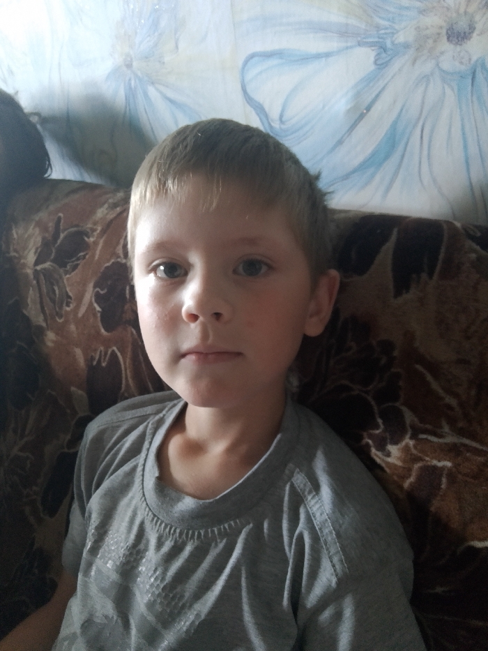 A young boy with blonde hair and blue eyes is sitting on a couch. He is wearing a gray t-shirt. The couch has a brown floral pattern. The background is a light-colored wall with a blue floral design. The image appears to be taken indoors. The location is identified as Изыхские Копи, Russia, which is a village in the Khakassia region.