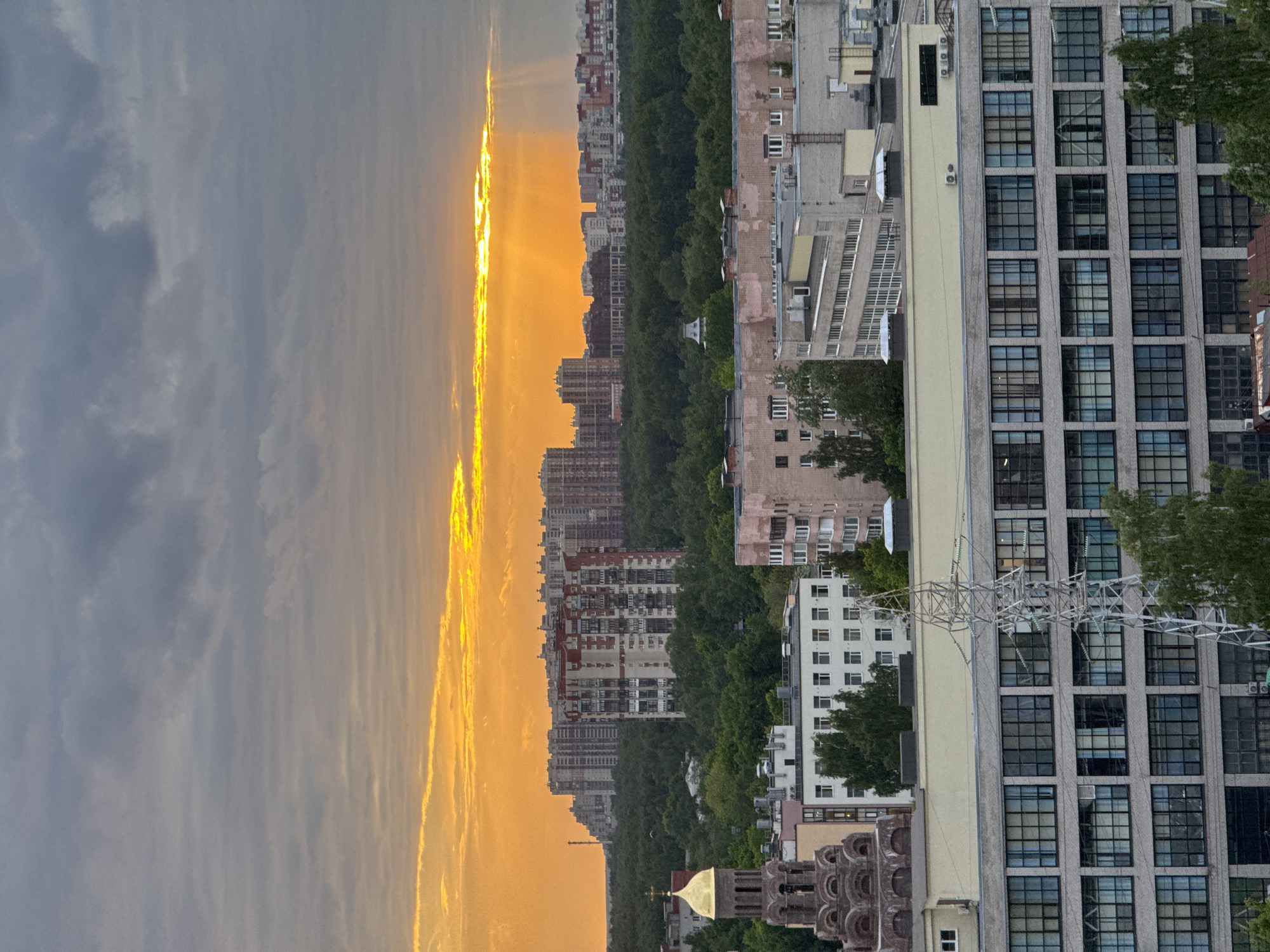 The image shows a cityscape at sunset in Unknown, Russia. The sky is a mix of dark clouds and bright orange light from the setting sun. A row of tall apartment buildings is visible in the distance, separated by a line of trees. In the foreground, there are more buildings, including a tall, modern building with many windows and a church with a golden dome. Power lines are visible in the lower right of the image.