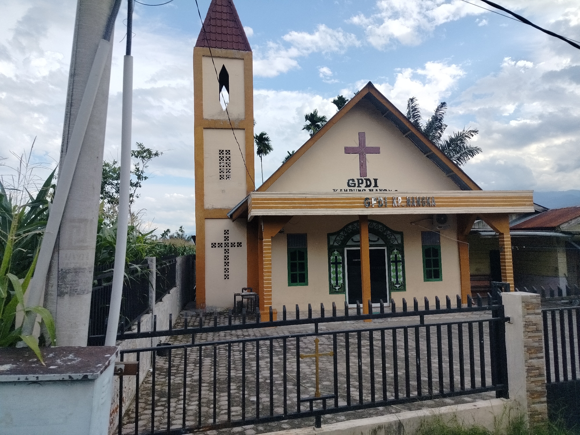 An exterior, eye-level, medium shot captures a small church building with a bell tower in Lawepolak, Indonesia. The church is painted a light beige with orange trim and features a pitched roof with a red tile covering. A prominent purple cross is affixed to the facade above the entrance. Text above the entrance reads "GPDI" and below that is "KP. MANGKA." The bell tower, also beige with orange trim, has a tall, narrow opening and decorative brickwork. The foreground is dominated by a black metal fence and a walkway paved with hexagonal stones. A small golden cross stands within the fenced area. To the left of the church, a tall structure with white pipes is visible, along with dense green vegetation, including what appears to be corn stalks. The sky is partly cloudy with white clouds against a blue background. The overall scene suggests a tranquil, perhaps rural, setting for a place of worship.