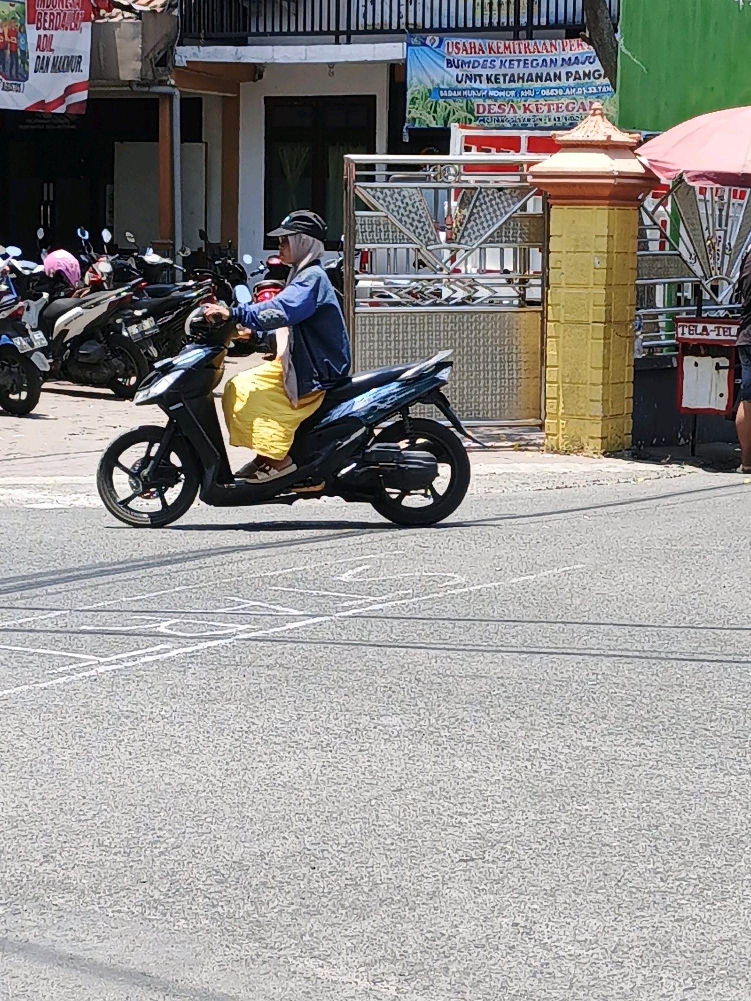 The image shows a street scene in Ketegan, Indonesia, on a sunny day. In the foreground, a person wearing a light blue jacket, grey headscarf, and yellow skirt rides a black and blue scooter. The person is wearing white sandals. Behind the scooter rider, several other motorcycles are parked. The background features a building with a white facade, a yellow pillar with decorative elements, and a red and white banner with Indonesian text. The street is paved with asphalt and marked with white lines. A pink umbrella is visible on the right side of the image.