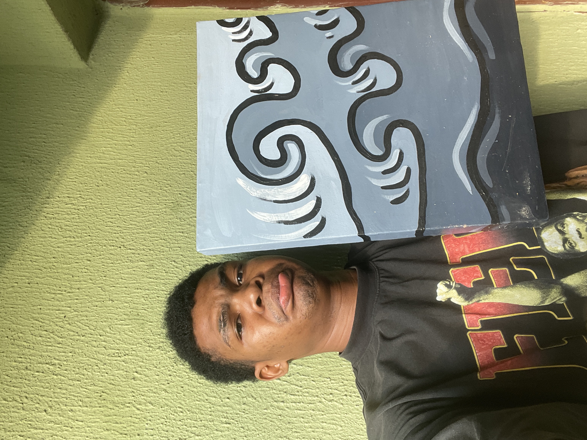 A man is holding a painting in front of a green wall. The painting depicts stylized waves in shades of gray and black. The man is wearing a black t-shirt with a large graphic. The location is outdoors, in Ado, Nigeria, as per the prompt.