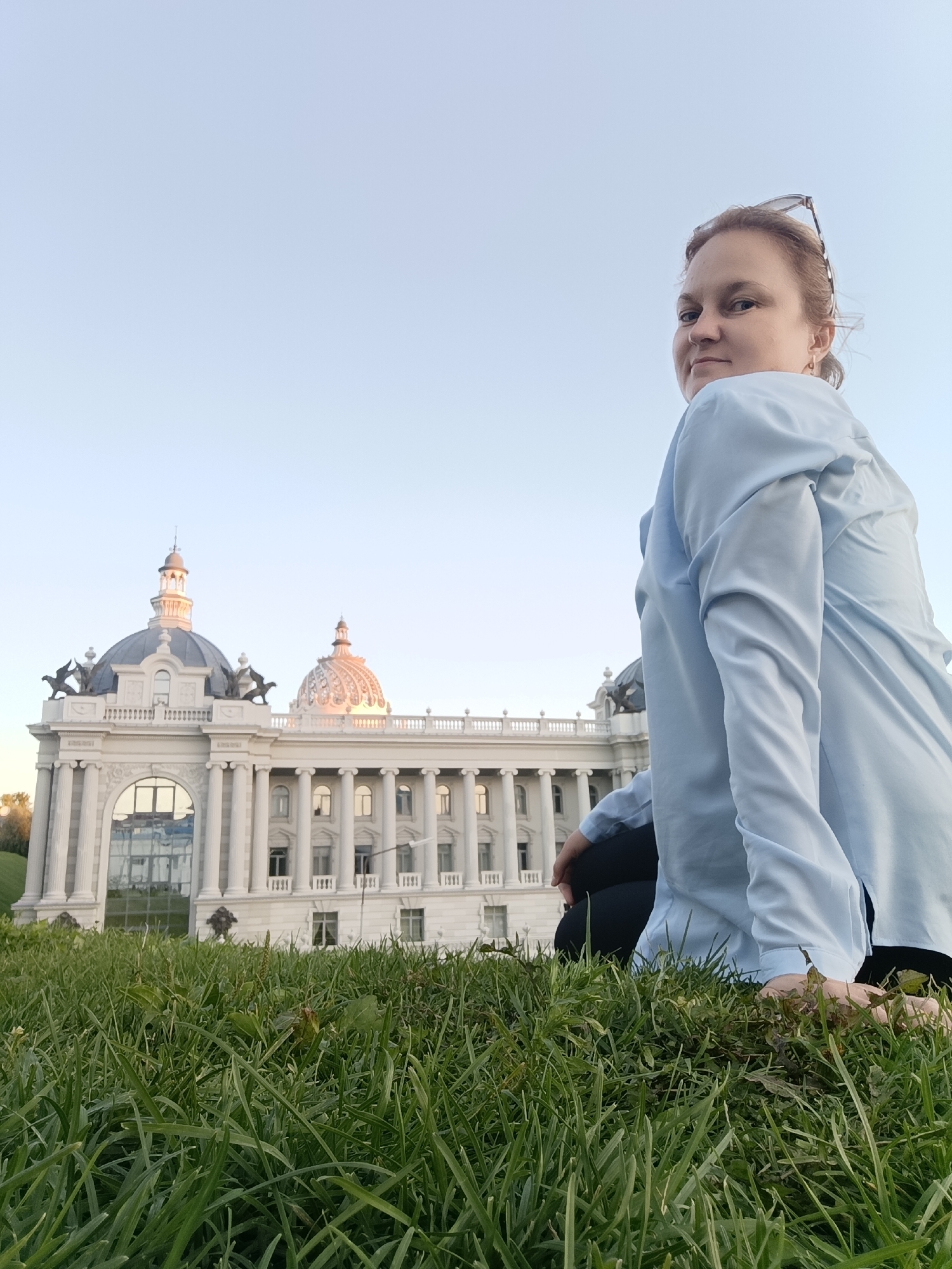 A woman in a light blue shirt and dark pants is sitting on a grassy slope in front of the Kazan Family Center, also known as the "Chalice." The building, a landmark in Kazan, Russia, features a distinctive dome and classical architecture with columns and ornate detailing. The woman is looking over her left shoulder towards the camera with a slight smile. Her sunglasses are pushed up on her head. The sky is clear and pale blue, and the lighting suggests it is late afternoon or early evening. The foreground is dominated by lush green grass, with the building and the woman in the background.