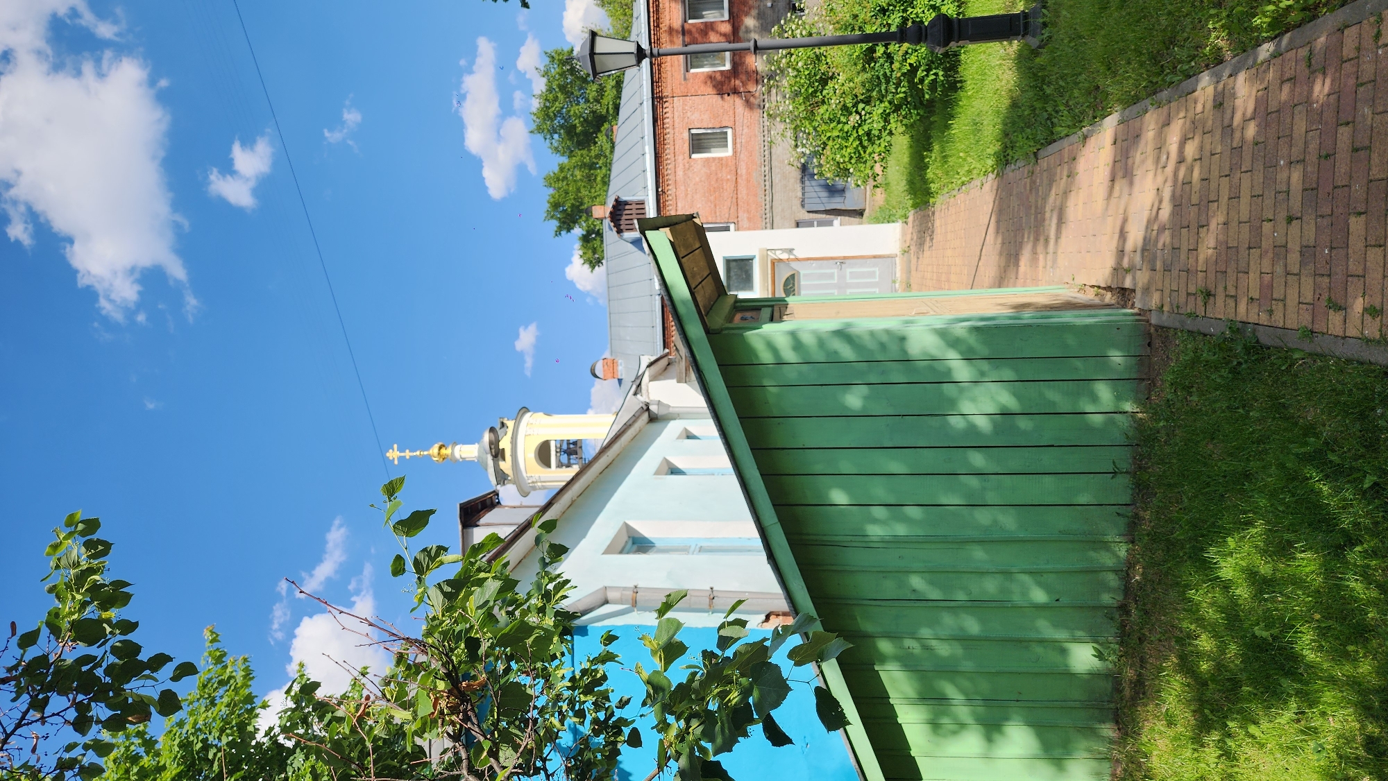 The image captures a bright, sunny day in what appears to be a historical urban setting in Moscow, Russia. The perspective is slightly upward-angled, showcasing a mix of architecture and natural elements under a vibrant blue sky.
In the foreground, lush green foliage with individual leaves is visible on the left side, partially obscuring the buildings behind it. A distinctive green wooden fence, constructed from horizontal planks and showing shadows of overhead leaves, runs diagonally across the mid-ground from the bottom right towards the center. To the right of the fence, a neatly paved pathway made of light brown and reddish bricks stretches into the distance, bordered by green grass.
In the mid-ground, a light blue building with white window frames and a blue foundation is prominent on the left. Above its roofline, the golden onion dome and spire of an Orthodox church, topped with a cross, gleam under the sun. Further back and to the right, a multi-story reddish-brown brick building with several windows and a gray roof is visible. A tall, black, ornate street lamp stands in front of this brick building. Between the brick building and the green fence, another white building with a gray door and window can be seen.
The background is dominated by a clear blue sky dotted with scattered white cumulus clouds. A thin, dark power line is faintly visible stretching across the sky. The time of day appears to be mid-morning or early afternoon, characterized by bright sunlight and well-defined shadows, indicating clear weather. No people or visible text are present in the scene.