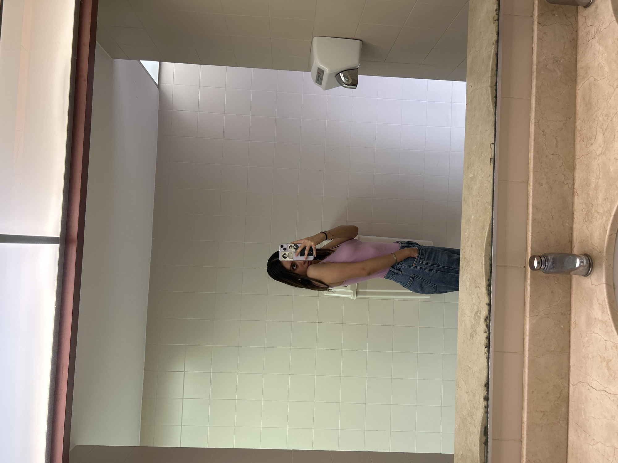 A photograph shows a reflection in a bathroom mirror. The reflection reveals a person taking a picture of themselves with a smartphone, wearing a pink top and jeans. The bathroom has white tiled walls, a white hand dryer, and a marble sink. The location is Unknown, Unknown.
