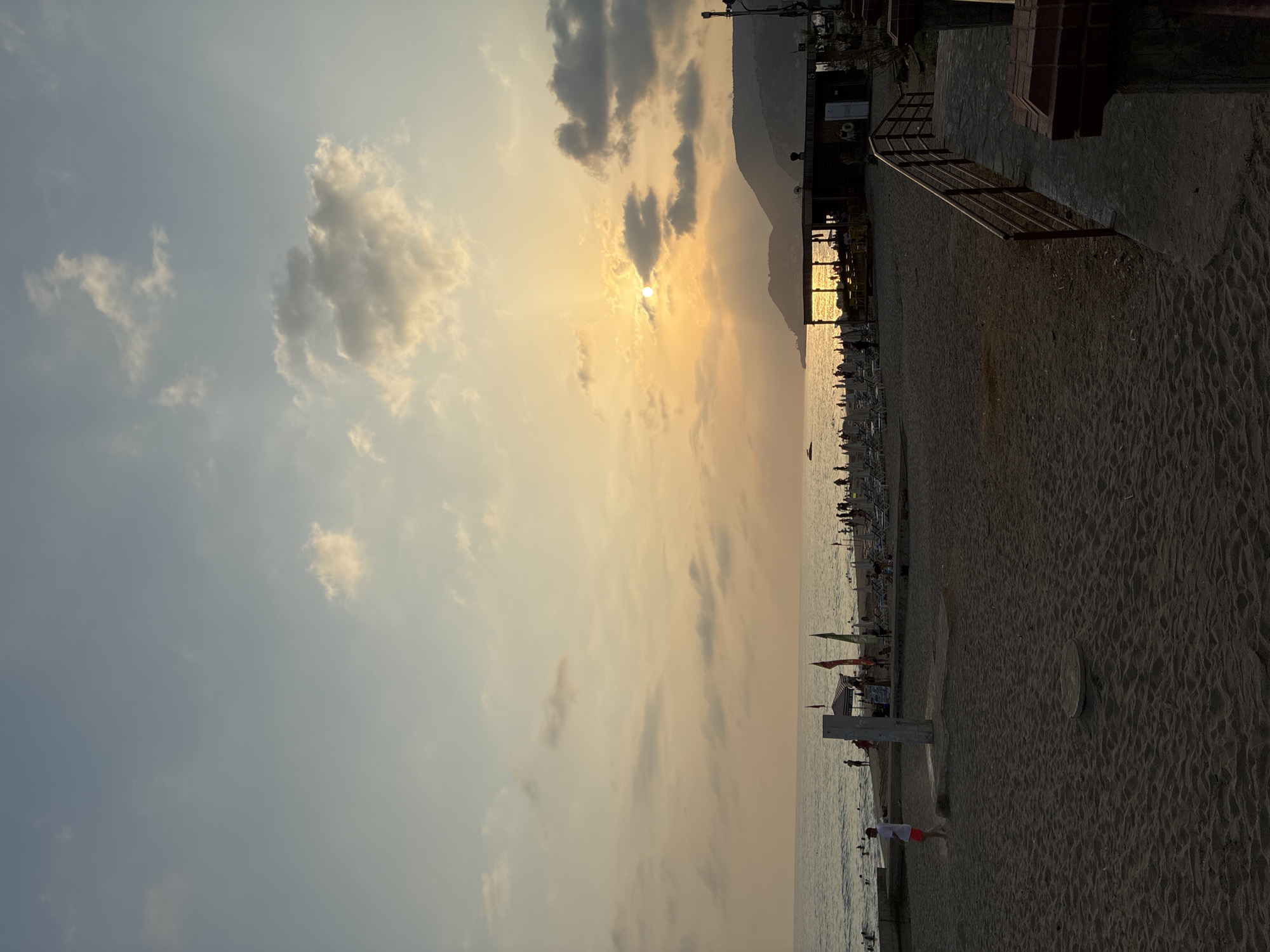 The image depicts a beach scene in Alanya, Turkey, during sunset. The sun is low in the sky, casting a warm glow over the water and the sandy beach. Several people are visible on the beach and in the water, enjoying the evening. Beach umbrellas and lounge chairs are set up, indicating a public beach area. A building, possibly a restaurant or beach bar, is situated on the sand, with people gathered around it. In the background, hills or mountains are silhouetted against the colorful sky. The sand is textured with footprints and the gentle waves of the sea are visible. The overall atmosphere is relaxed and serene, typical of a beach at dusk.