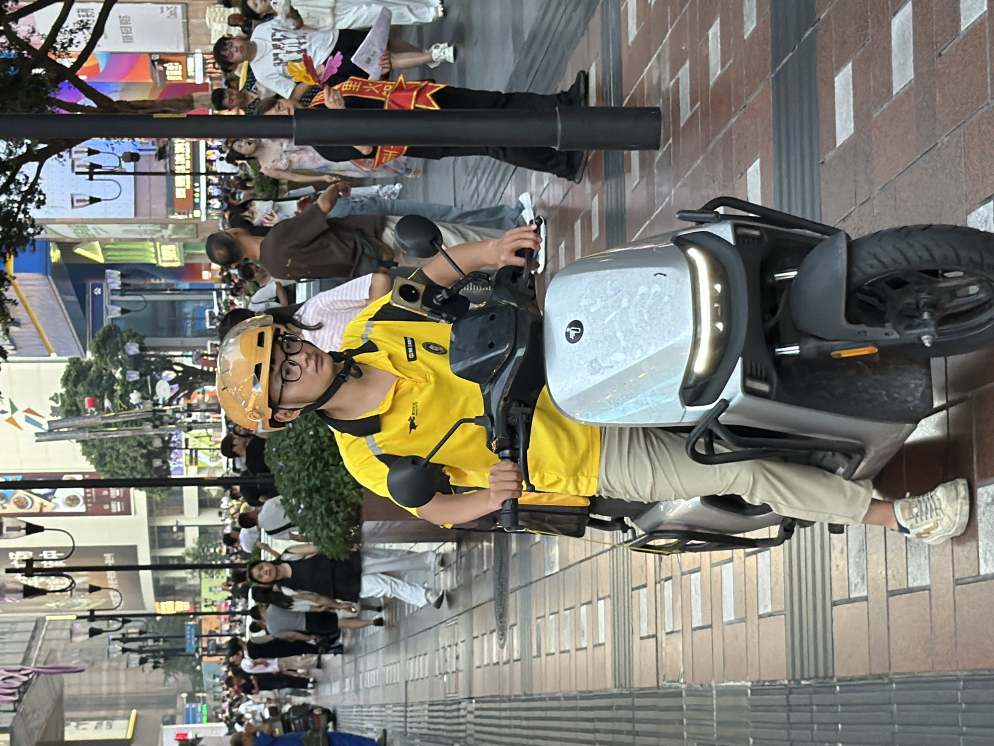 The image depicts a street scene in Yuzhong District, China, likely in a pedestrian-heavy commercial area. In the foreground, a young man wearing a bright yellow polo shirt with black accents and a yellow helmet is seated on a modern-looking grey electric scooter. He is wearing khaki pants and white sneakers. His right hand is on the scooter's handlebars. The scooter has a distinctive front design with illuminated accents. The background shows a bustling street with numerous pedestrians walking on a paved sidewalk. Buildings with signage and some greenery are visible. The overall lighting suggests it is daytime, possibly late afternoon or early evening.