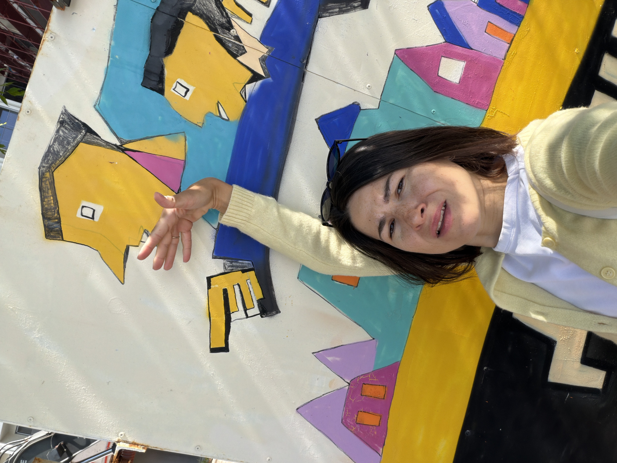 A woman is posing for a selfie in front of a large, colorful abstract mural. She has short, dark hair and is wearing a light yellow cardigan over a white shirt. Sunglasses are perched on her head. Her right arm is extended, with her hand gesturing towards the left side of the mural.
The mural features stylized, geometric depictions of people and buildings in bright colors like yellow, blue, purple, and orange. The background of the mural is white.
The location is likely in Rafailovići, Montenegro, based on the user's request. However, without additional visual cues in the image, this cannot be definitively confirmed from the photograph alone. The picture appears to be taken outdoors in natural light.