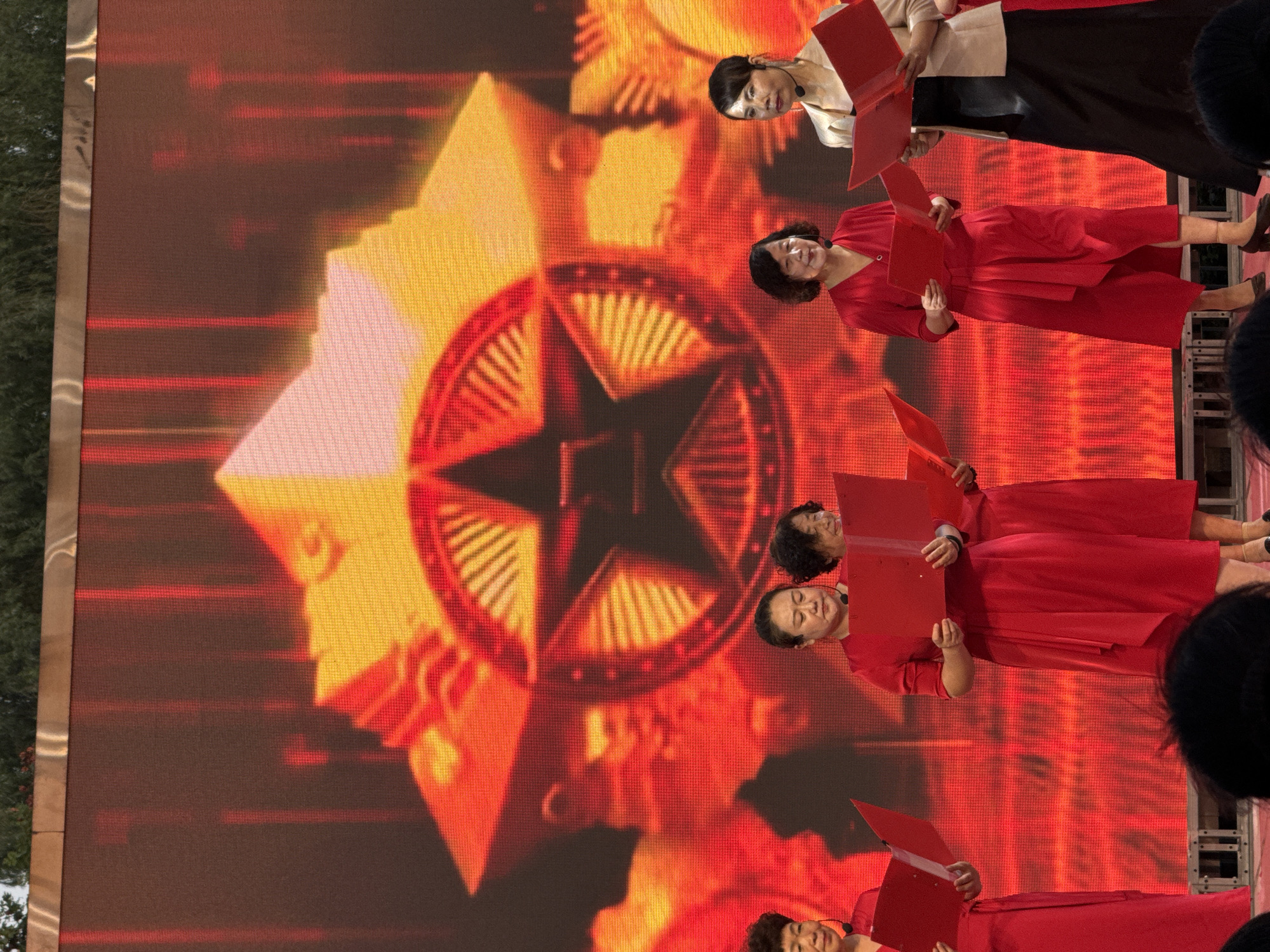 The image shows a group of women performing on a stage in front of a large LED screen displaying a red and yellow star emblem. Five women are visible, all holding red folders, suggesting they are singing or reciting. They are dressed in red dresses, except for the woman on the far right, who is wearing a tan jacket over a black dress. The background screen is illuminated with bright red and yellow lights, creating a dramatic effect. The stage appears to be set up for an outdoor event, as indicated by the foliage visible at the top of the image. The overall scene suggests a formal or celebratory performance. Based on the available information, this event is likely located in Jinzhou, China.
