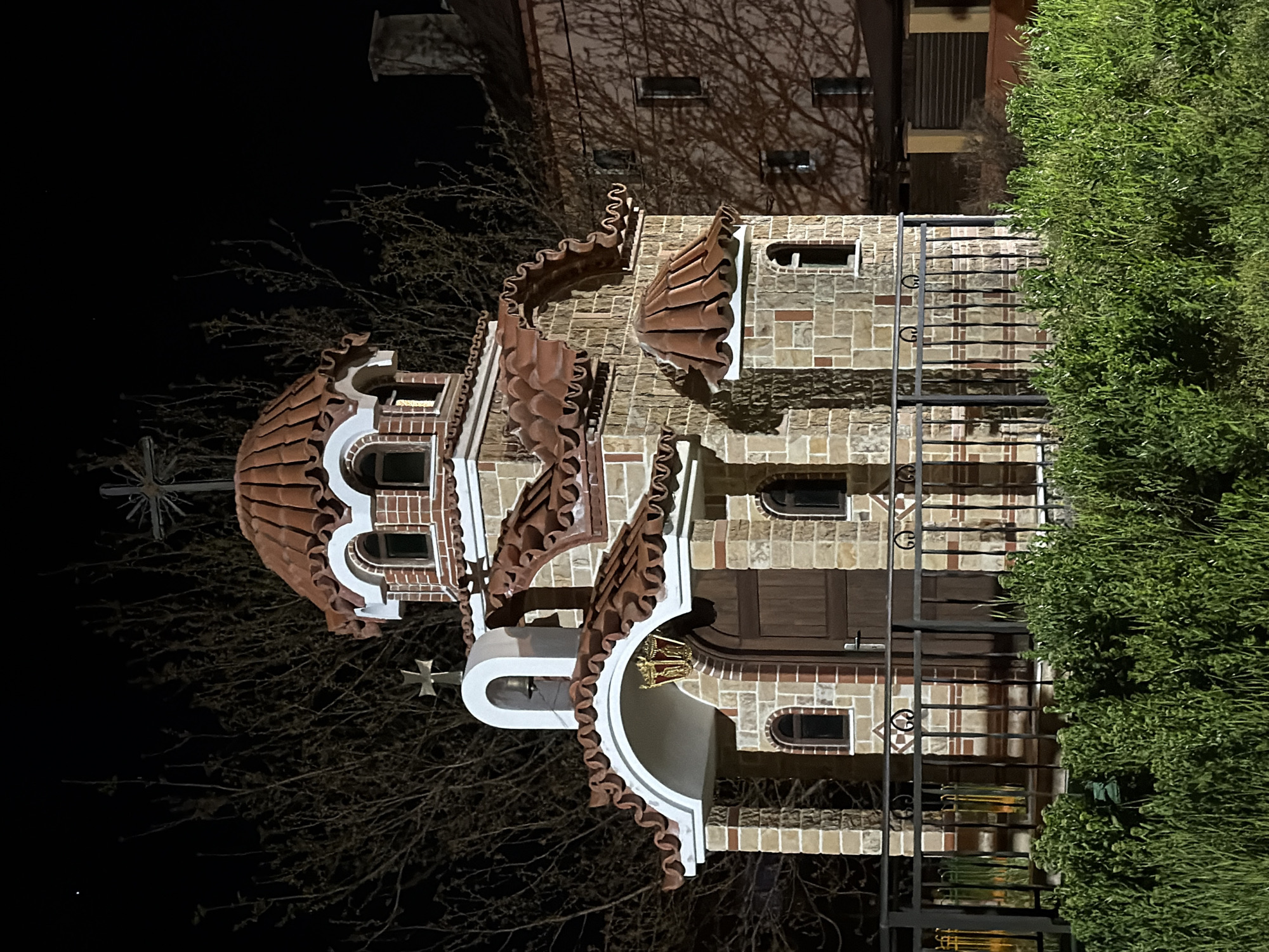 The image depicts a small church at night, identified as the Russian Church in Sofia, Bulgaria, due to its distinctive architectural features. The church has a brick facade with decorative red tile work around the roof and windows. A wrought-iron fence is visible on the right side of the frame. The lighting is artificial, illuminating the church. The top of the structure includes a dome. Parts of bare branches are next to the church. A green hedge is partially visible on the right, and a building is in the background. The image is taken from an eye-level perspective.