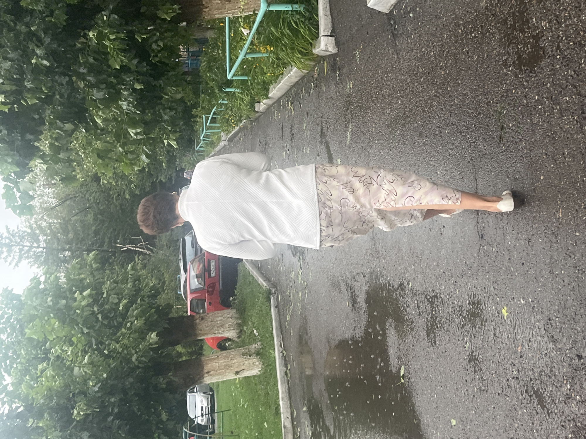 A woman with short, dark hair is walking away from the camera down a wet asphalt path. She is wearing a white blazer with a textured pattern and a light-colored skirt with a swirling design. She has on white slip-on shoes. The path is lined with trees on the left and a grassy area with parked cars, including a red hatchback, on the left. On the right, there is a green metal railing, a strip of grass with yellow flowers, and more trees. The asphalt is dark and reflective due to recent rain, with puddles and scattered leaves. The sky appears overcast. The location is the city of Oskemen, Kazakhstan.