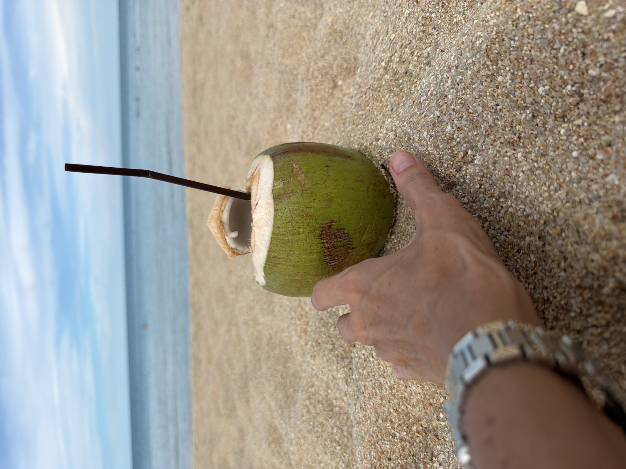 A person's hand, wearing a metallic watch on the left wrist, reaches towards a green coconut with a straw in it. The coconut rests on a sandy beach. In the background, the ocean meets the sky under a cloudy blue sky. The scene is likely located in Baan Chaweng, Thailand.