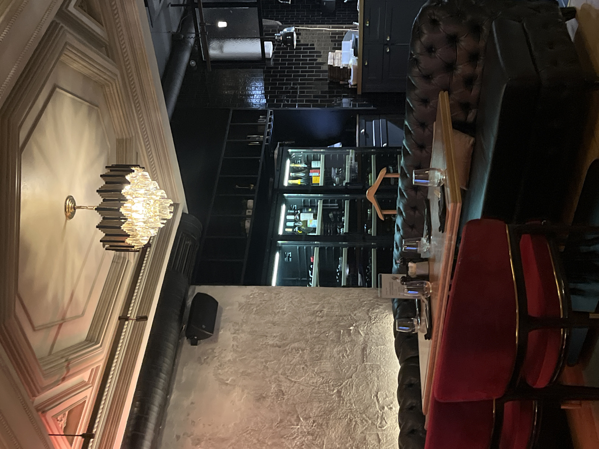An interior view of an unoccupied restaurant or bar is presented, likely in the city of Unknown, Unknown. The scene features a mix of classic and industrial design elements under warm, artificial lighting.
The ceiling is ornate, with a multi-layered coffered design in a light, possibly cream, color. A prominent, multi-tiered crystal chandelier with a brass-colored metal frame hangs centrally from the ceiling, illuminated and casting reflections. To the left, several dark, slender pendant lights are suspended. Exposed dark ventilation pipes run along the top left wall.
The left wall has a textured, light-colored finish, possibly stucco or concrete, with upward-facing lights illuminating its surface. A dark, wedge-shaped speaker is mounted high on this wall. The background on the right is dominated by dark-colored cabinetry and walls. Two large, glass-fronted wine or beverage refrigerators with glowing interiors display numerous bottles of wine and spirits. Above these units and extending to the right are dark shelving units and closed cabinets. A wooden coat hanger is visible hanging between the refrigerators and the central dark cabinetry.
Further to the right, a section of the wall is clad in dark, rectangular subway tiles. Below this, a dark counter area is equipped with more dark cabinetry. Stacks of white objects, possibly plates or napkins, are visible on the counter. An industrial-style pendant light and a dark, articulated desk lamp are present in this area.
In the foreground and midground, seating consists of a long, dark brown or black leather Chesterfield-style banquette, running along the tables. Two red velvet chairs with dark frames and subtle gold accents are positioned at a light-colored wooden table in the immediate foreground. The tables are set with clear water glasses, dark folded napkins, and condiment shakers (salt and pepper). No individuals are present in the image.