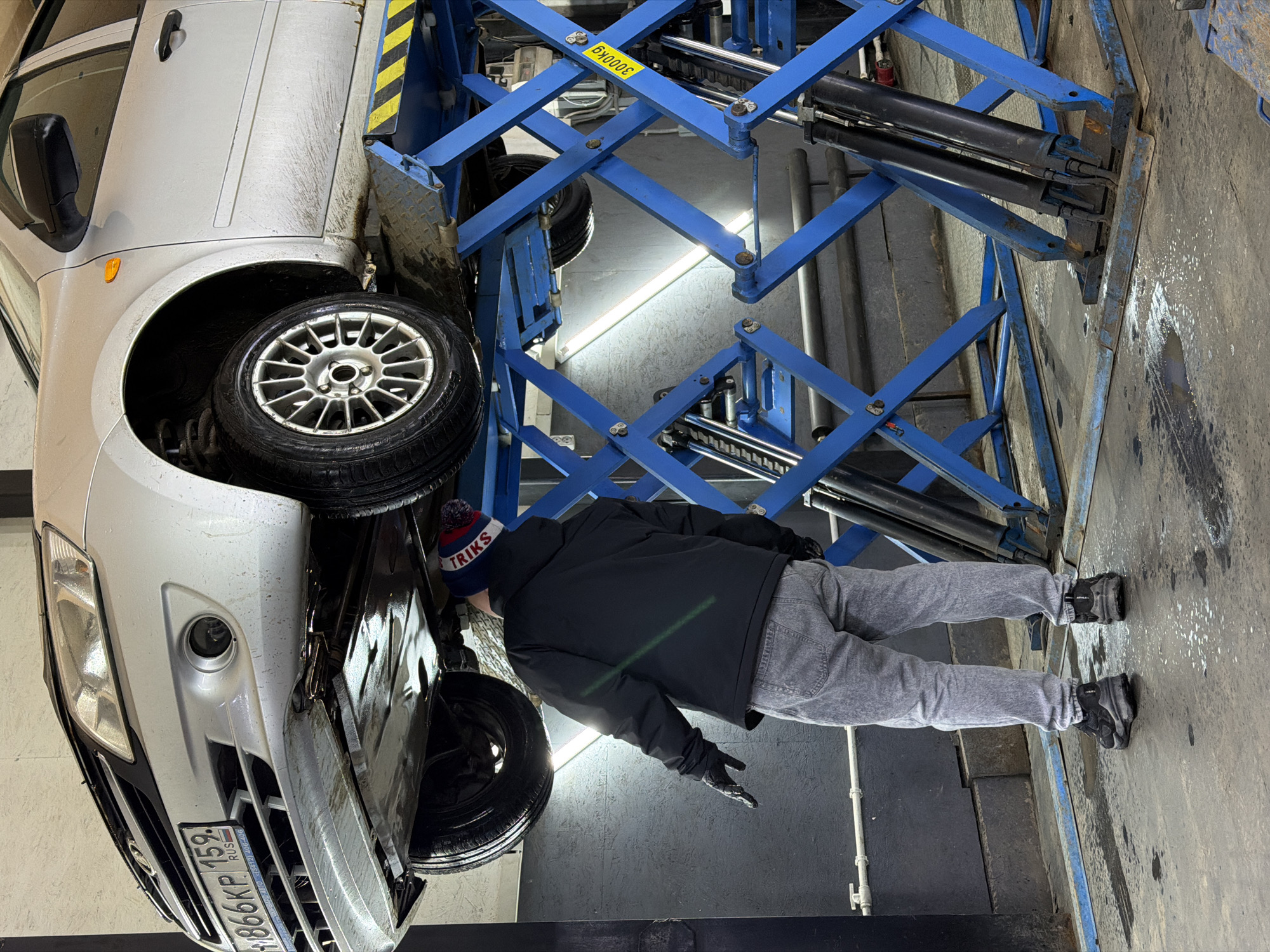 An indoor scene in what appears to be an automotive workshop or garage in Chaikovsky, Russia, shows a silver/light grey car elevated on a blue hydraulic scissor lift. The car, which looks somewhat dirty, has its front end raised, exposing its undercarriage and front wheel assembly.
A person, likely a mechanic, is standing beneath the elevated car, looking upwards as if inspecting or working on it. The individual is wearing a black jacket, grey jeans, black gloves, and a knit beanie with red, white, and blue stripes. The beanie has the word "TRIKS" visible on it.
The car lift is a robust blue structure, featuring multiple interconnected arms and hydraulic pistons. A yellow sticker on one of the lift's beams clearly indicates its capacity: "3000KG". The floor is concrete, and industrial lighting (a bright fluorescent tube) illuminates the area.
The car's front license plate is visible, displaying the alphanumeric sequence "M866KP 159 RUS", which corresponds to a Russian Federation license plate, with "159" being the region code for Perm Krai, where Chaikovsky is located. The activity taking place is car maintenance or repair within a professional garage setting. The overall impression is one of focused work in a functional environment.