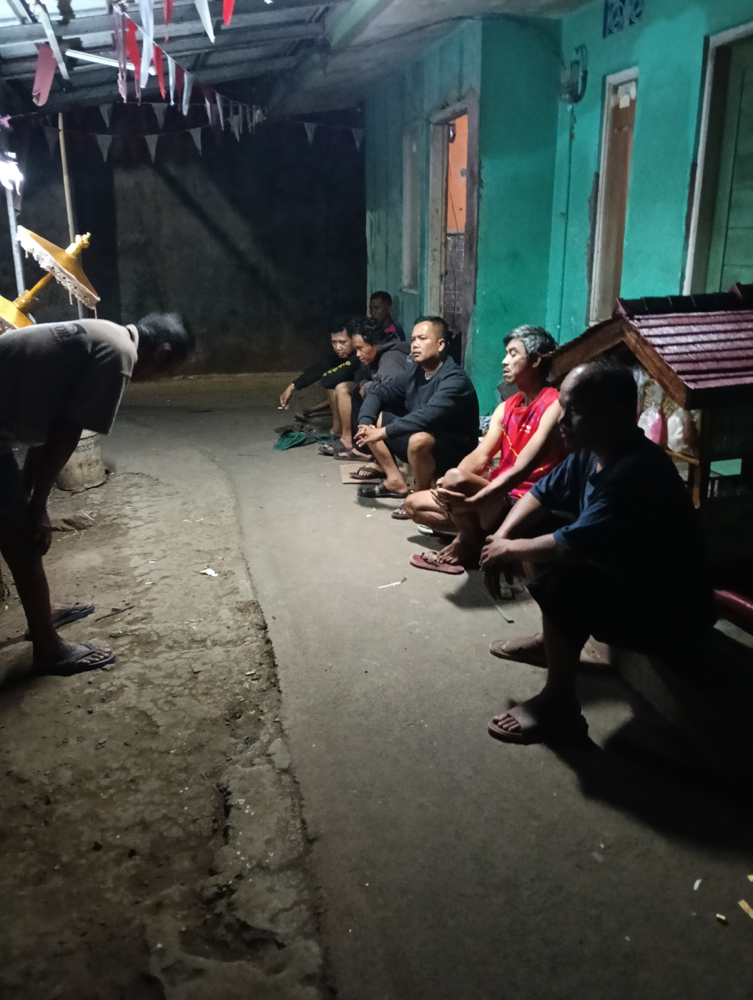 In Tasikmalaya, Indonesia, a group of men are gathered outdoors at night, illuminated by artificial lights and strung decorations. The scene appears to be in front of a building with a teal-colored facade and wooden window frames. Some men are sitting on the ground, while others are standing or crouching. They are dressed casually in t-shirts and shorts, with one man wearing a red sleeveless shirt. One man in the foreground is bent over, his face obscured. There is a small wooden structure with a tiered roof to the right of the frame. The ground is a mix of concrete and dirt, with scattered debris. The atmosphere suggests a community gathering or informal meeting in a residential area.